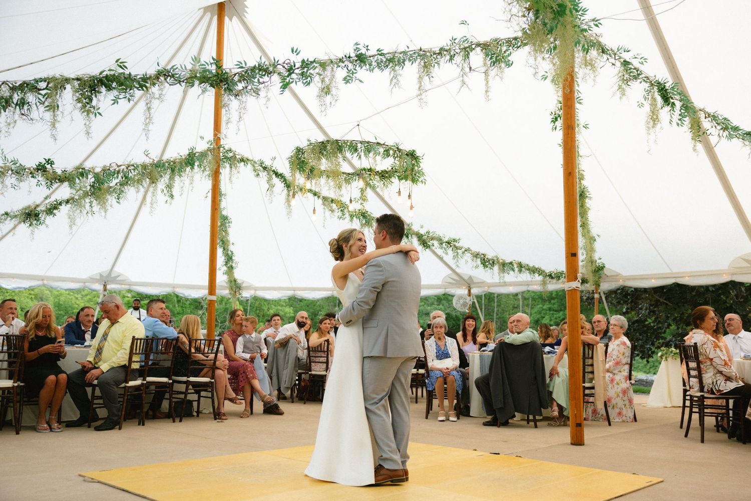 Bride and groom share first dance under elegant tent adorned with lush greenery garlands at outdoor wedding reception.