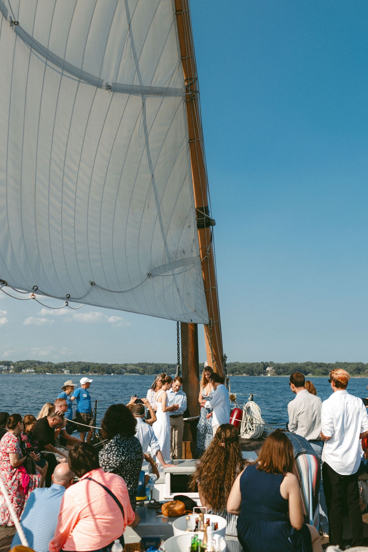 Guests gather on a sailboat deck during a celebration with white sails and blue water in view.