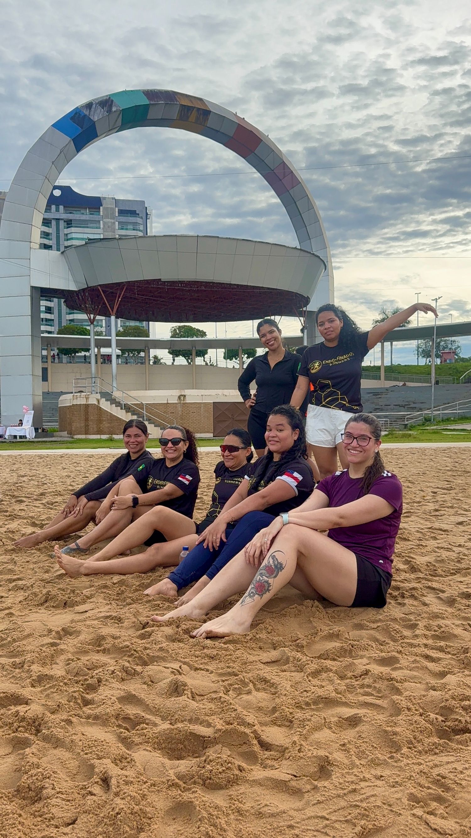 Group of female athletes posing on sandy beach in front of large circular architectural landmark.