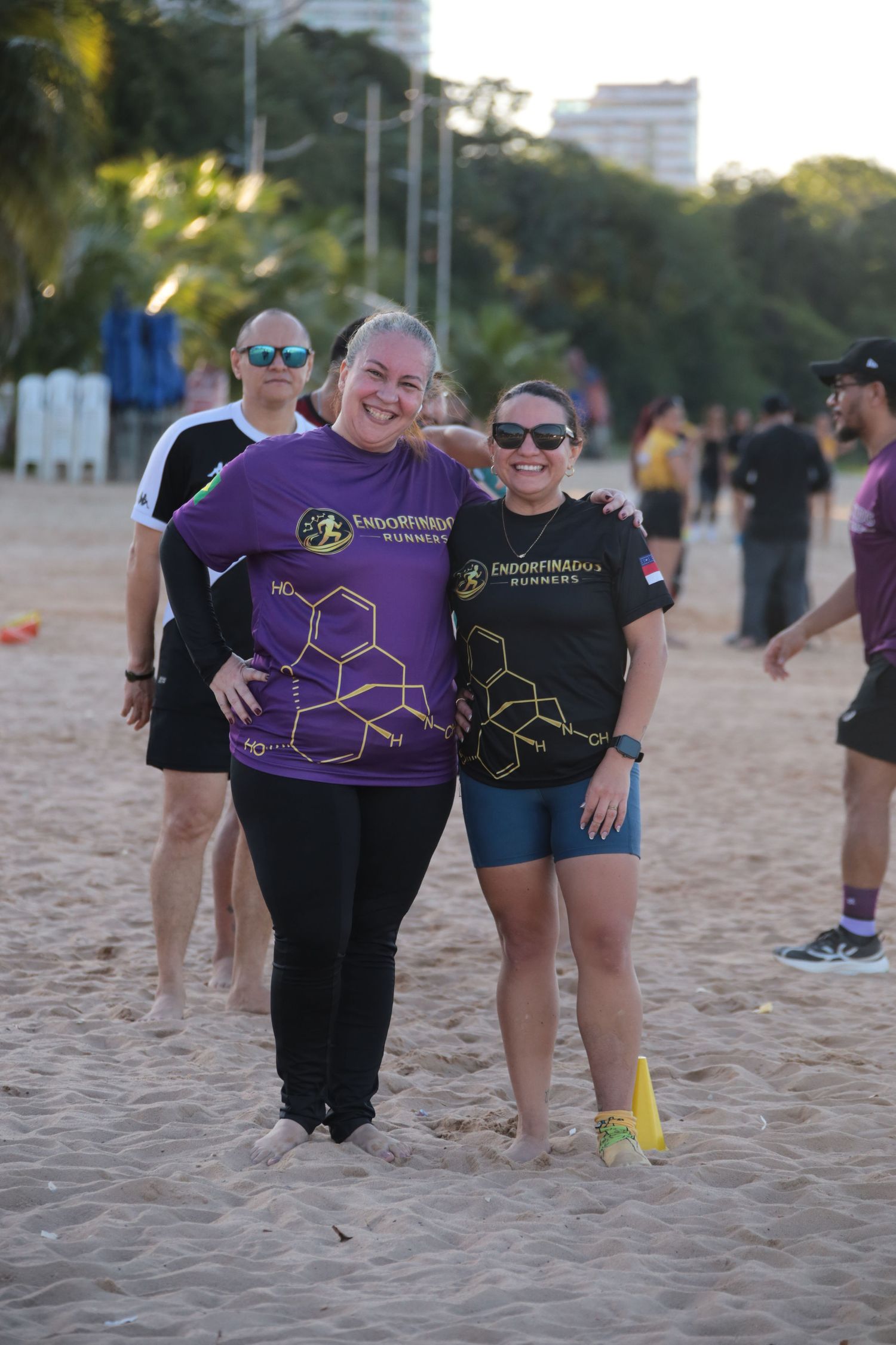 Two women in Endorfinados Runners shirts smiling together on a sandy beach at sunset.