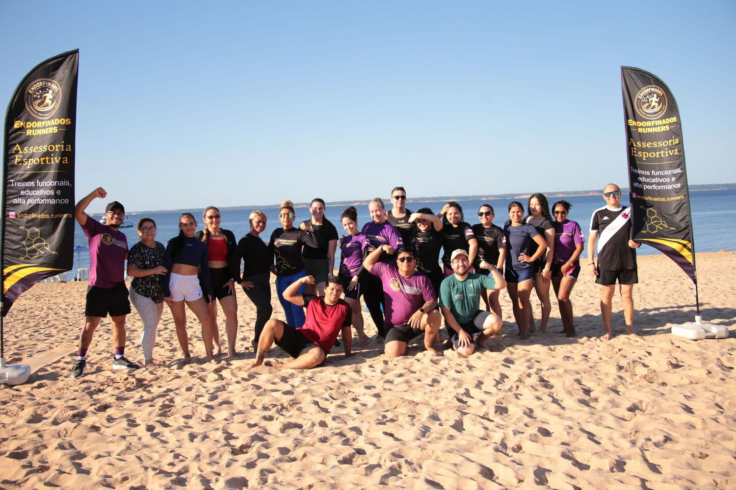 Large fitness group posing on a sunny beach with Assessoria Esportiva banners behind them.