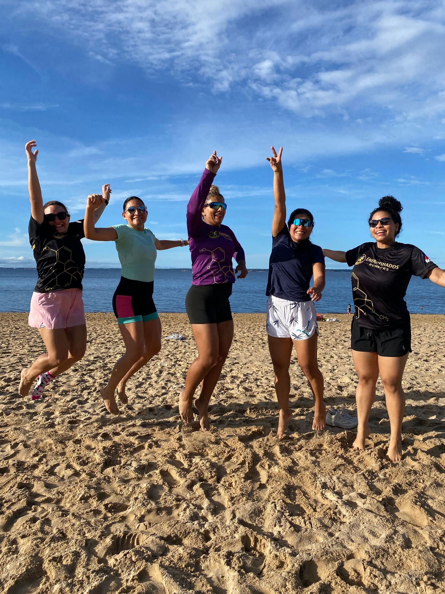Five athletic women jumping joyfully on a sunny beach with blue sky background.