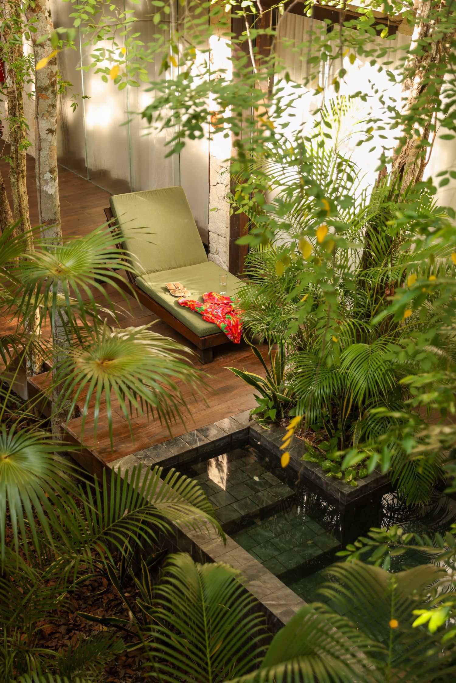 Lush tropical garden retreat with a green lounge chair on wooden deck beside a small reflecting pool.