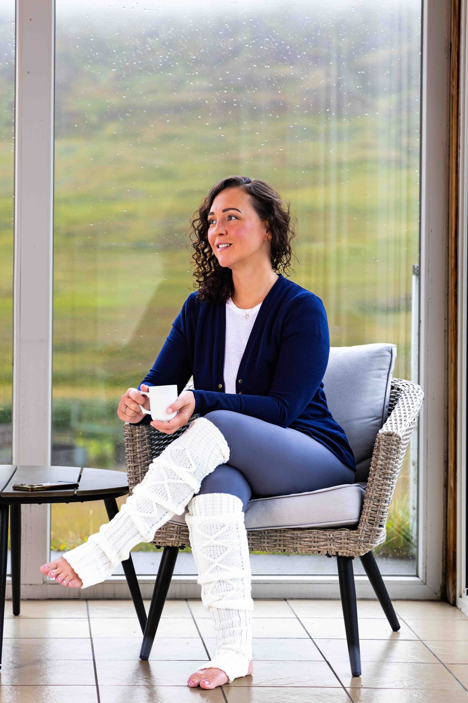 Woman in navy cardigan and white leg warmers sitting in wicker chair holding coffee mug by large window.