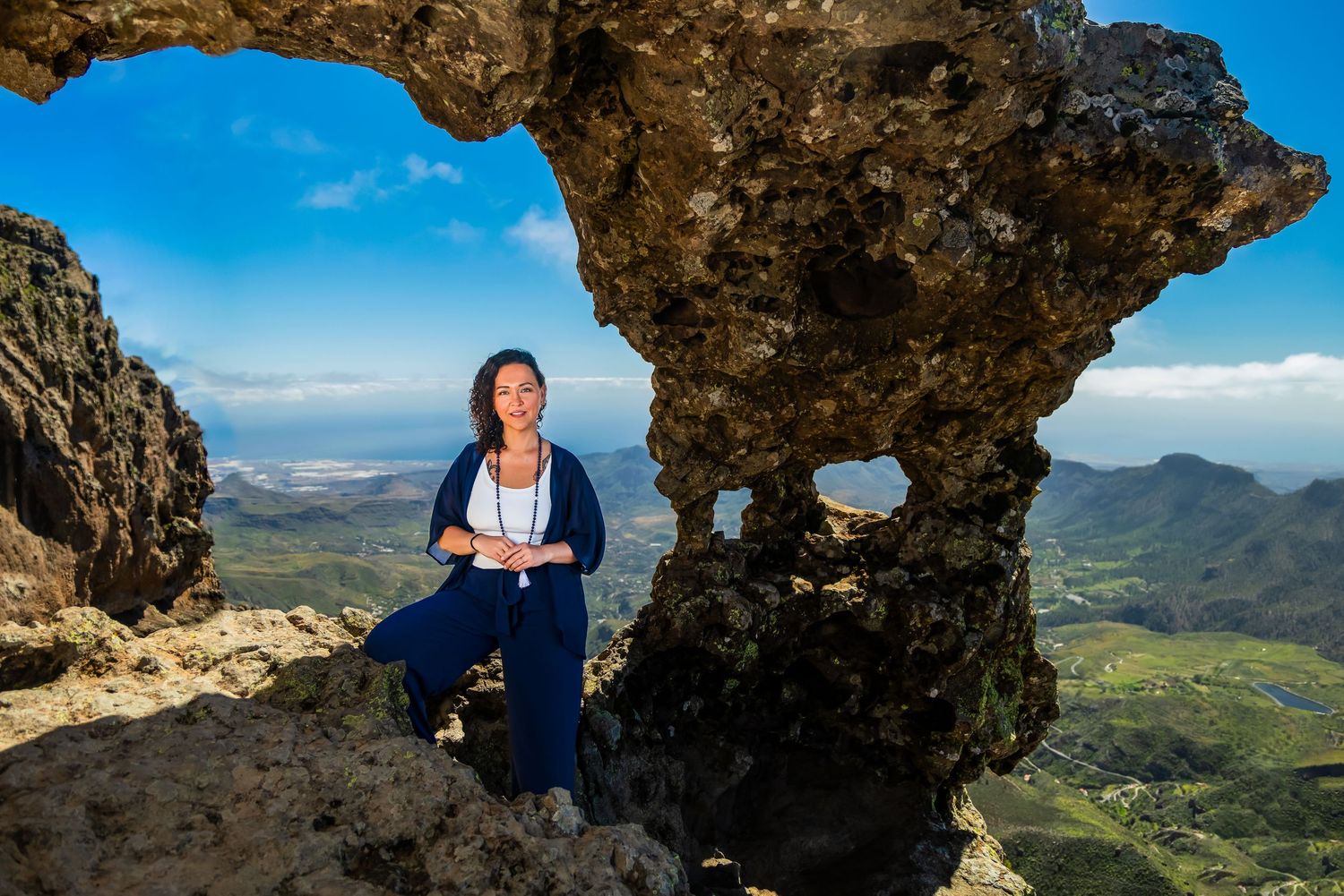 Woman sitting among dramatic rock formations on a mountain summit with panoramic valley and coastal views under blue sky.