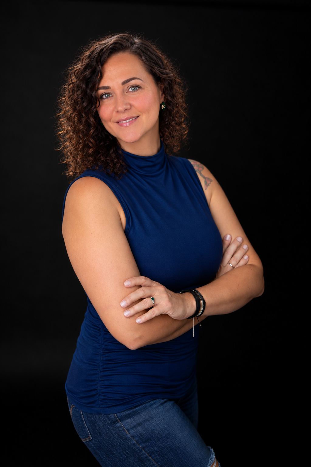 Woman with curly hair in blue sleeveless turtleneck and jeans poses confidently with arms crossed on dark background.
