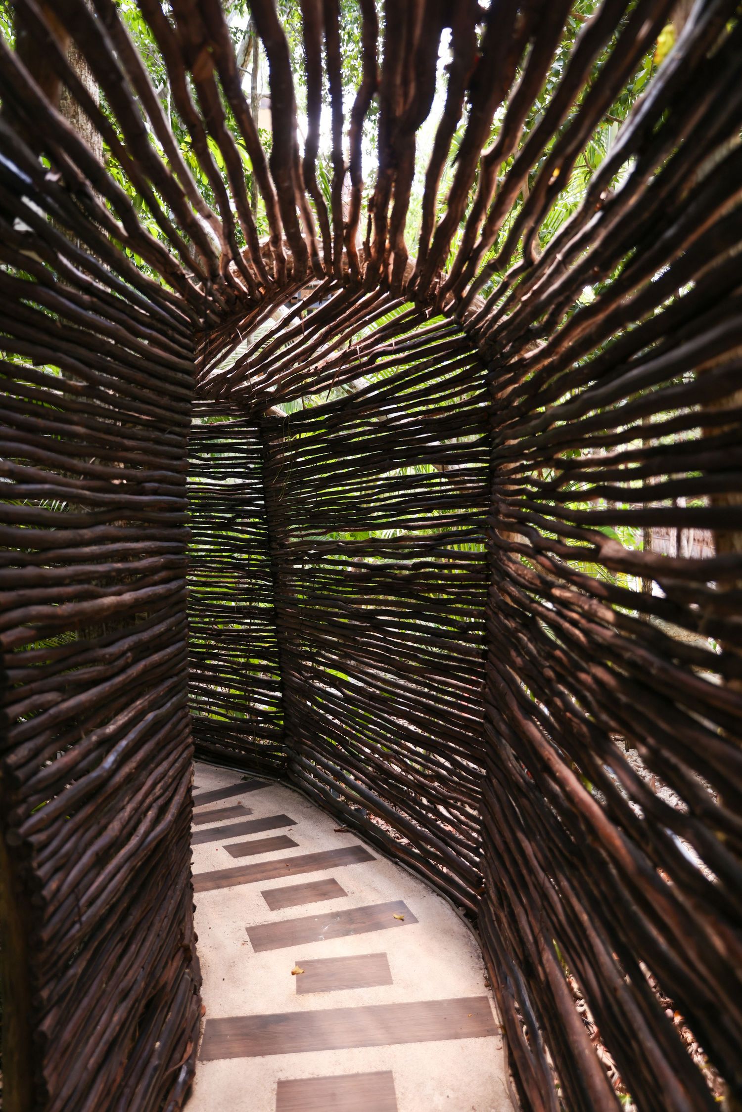 Rustic woven branch tunnel pathway with arched ceiling and tiled floor in a lush garden setting.