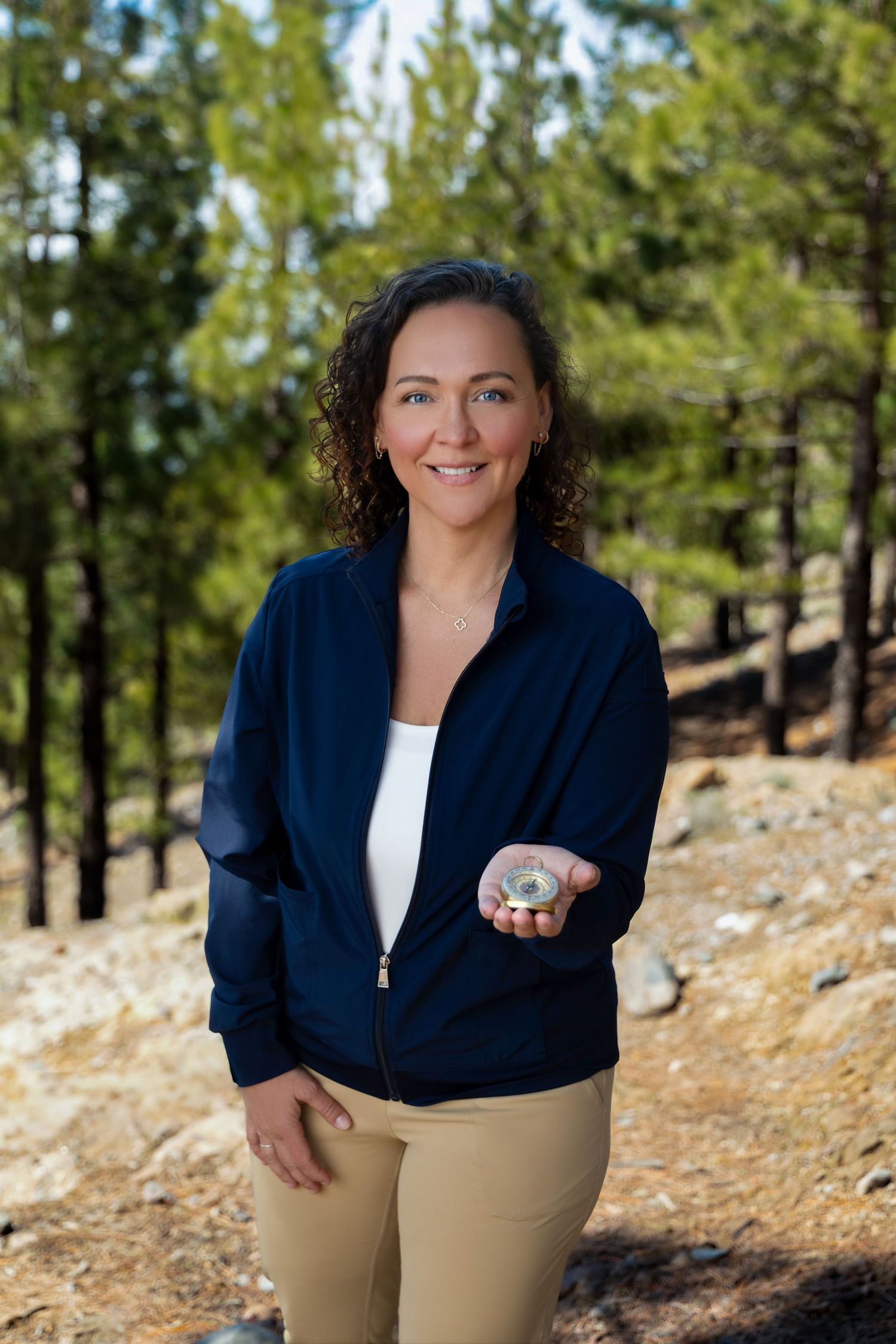 Woman in navy jacket holding a compass in a forest setting, smiling outdoors among pine trees.