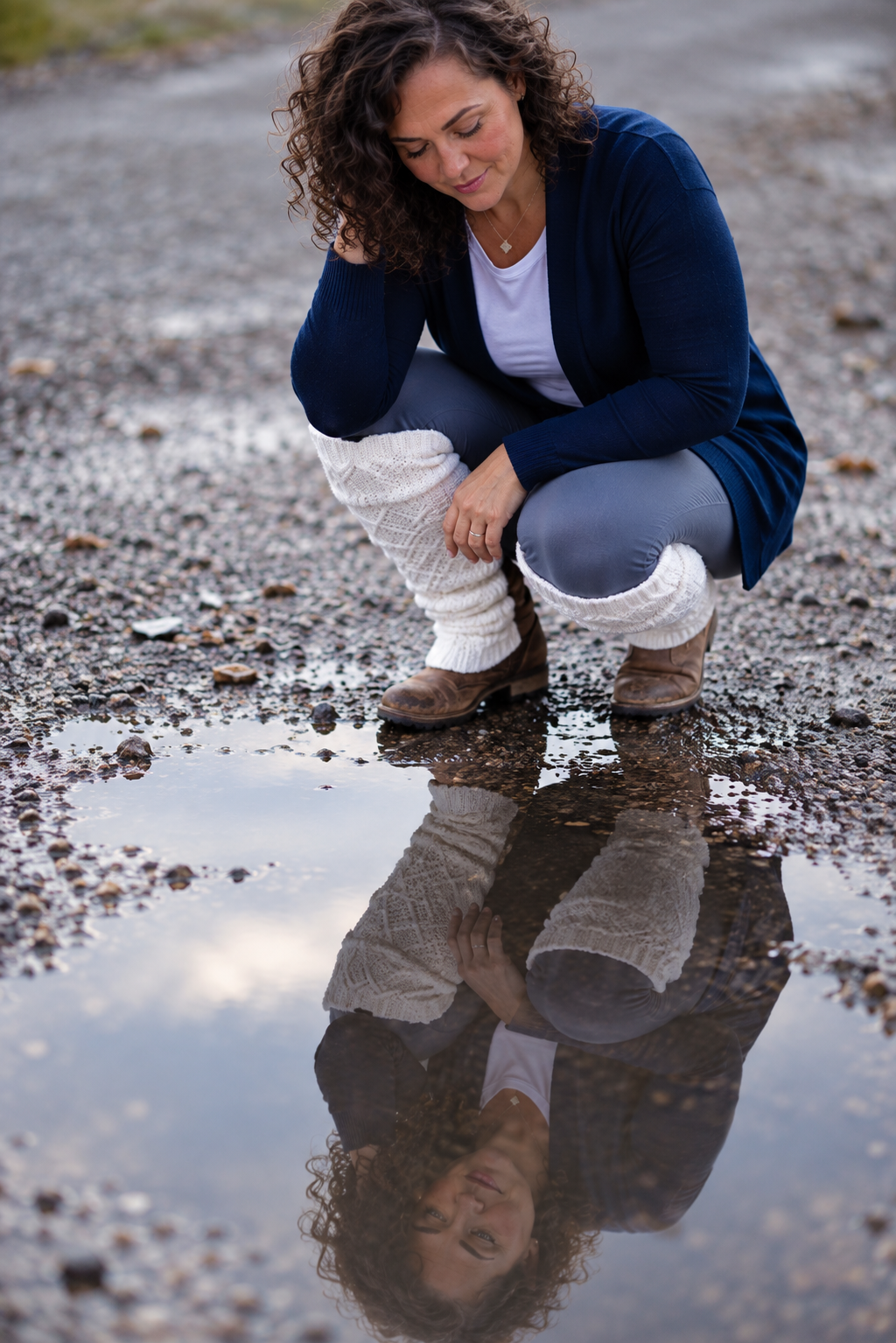Woman crouching beside a puddle on gravel, gazing at her reflection in the water, wearing a blue cardigan and white leg warmers.