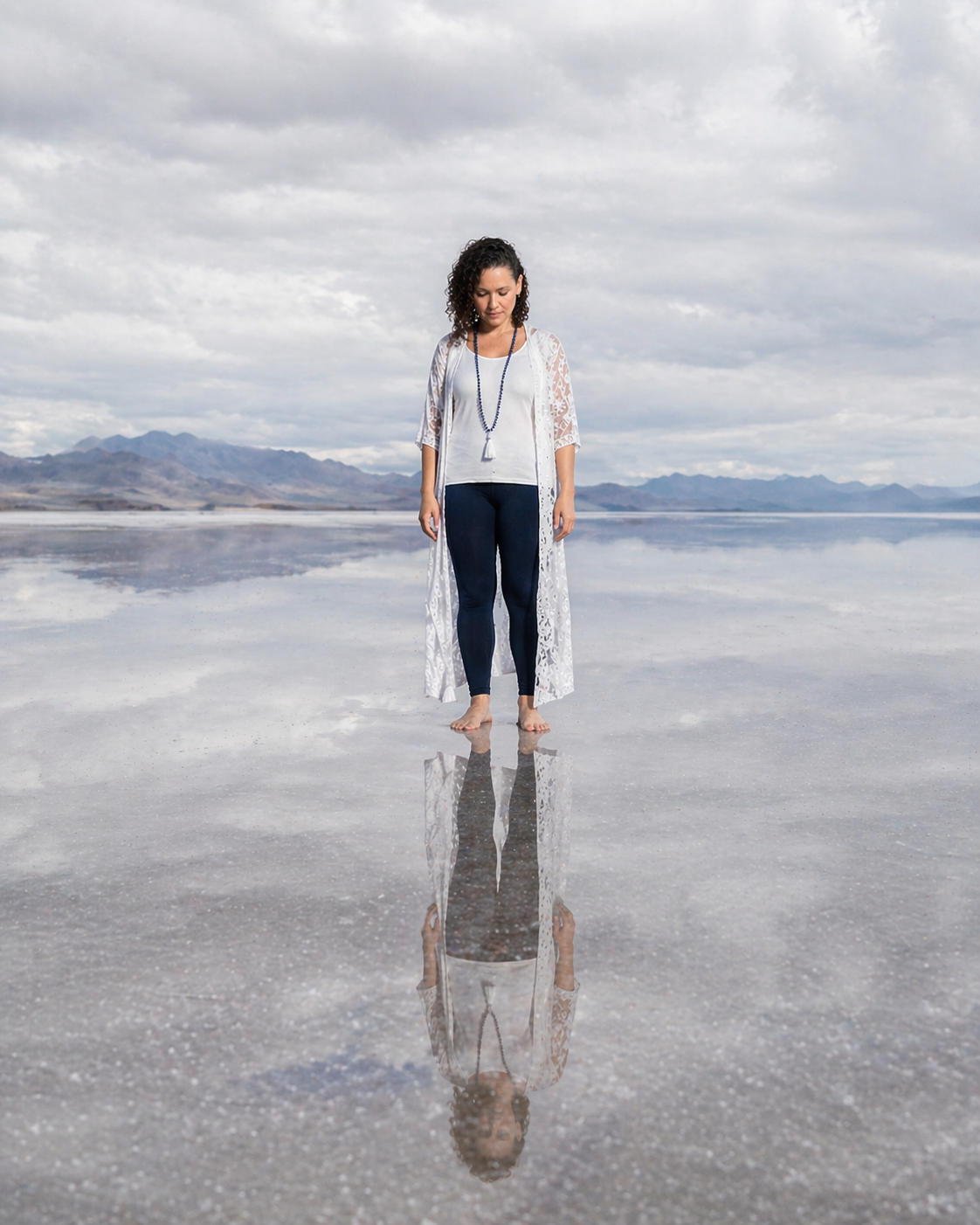 Woman standing barefoot on reflective salt flats with mountain backdrop and cloudy sky, wearing white lace cardigan and navy leggings.