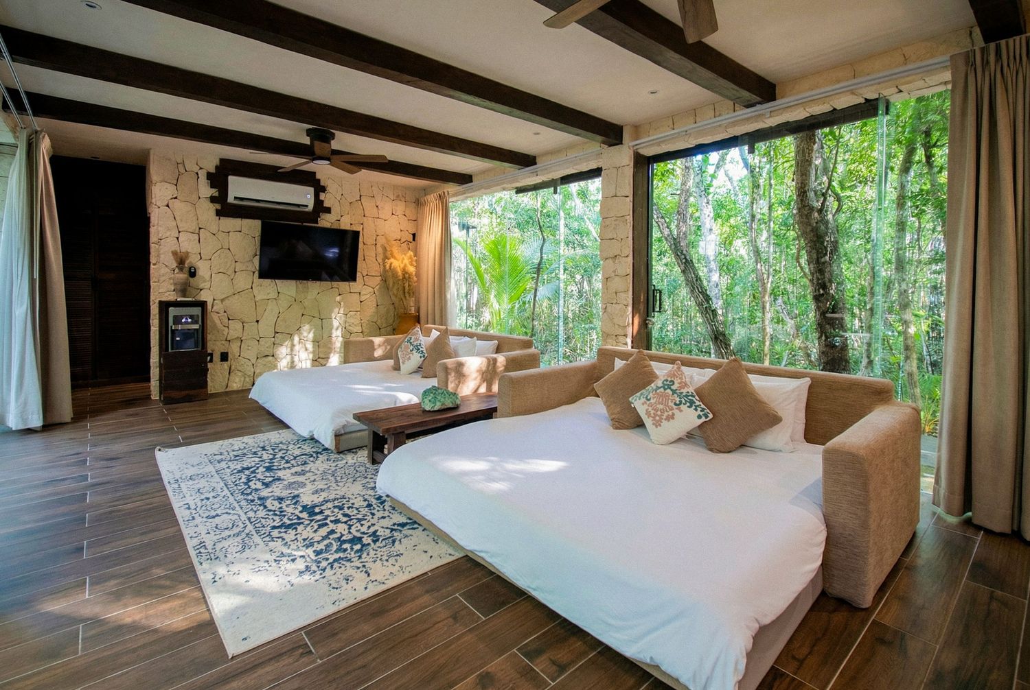 Bright jungle villa bedroom with two white beds, wood-beam ceiling, large windows overlooking lush green forest, and rustic stone wall.