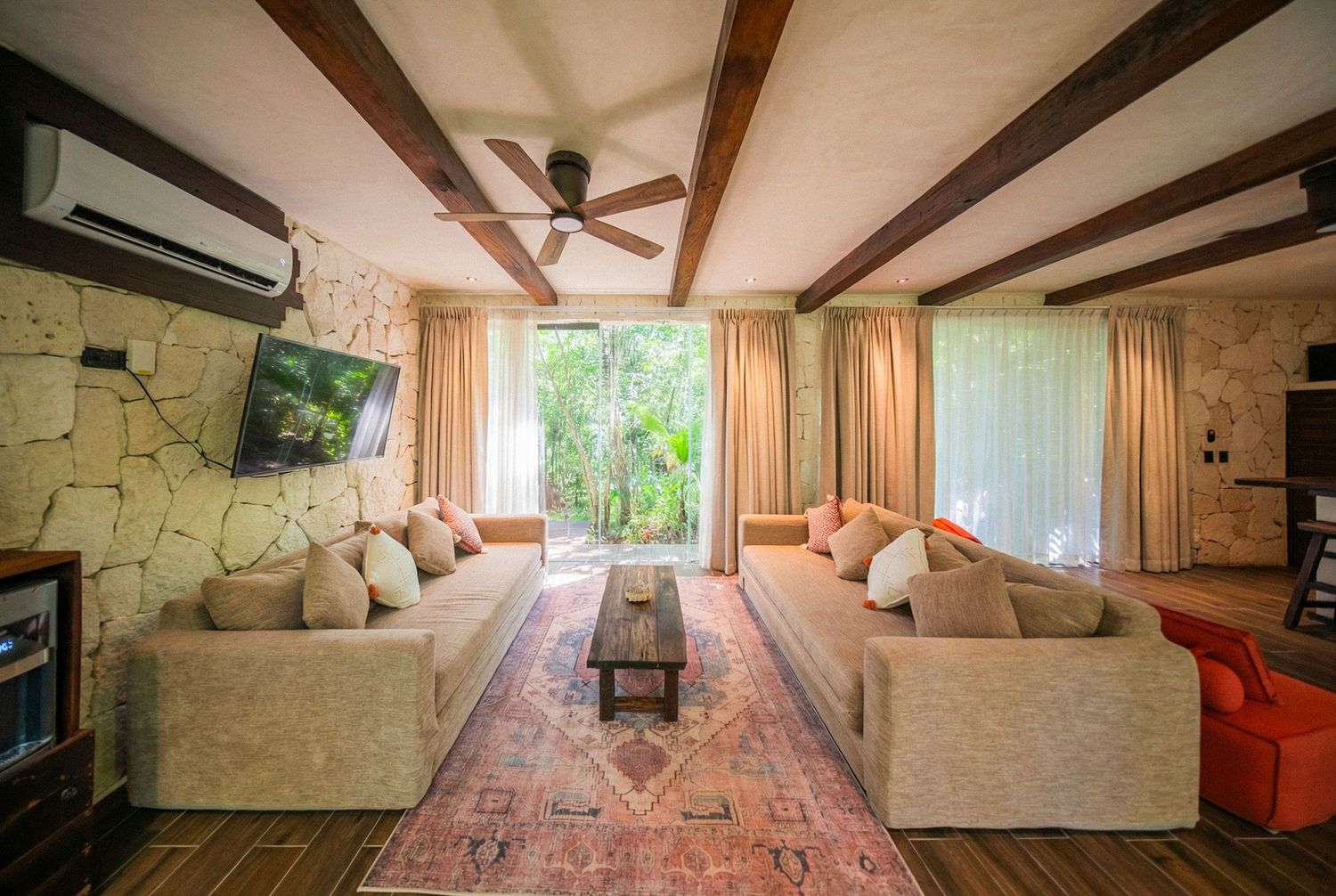 Spacious living room with beige sofas, wooden ceiling beams, ceiling fan, stone wall, TV, and lush garden view.