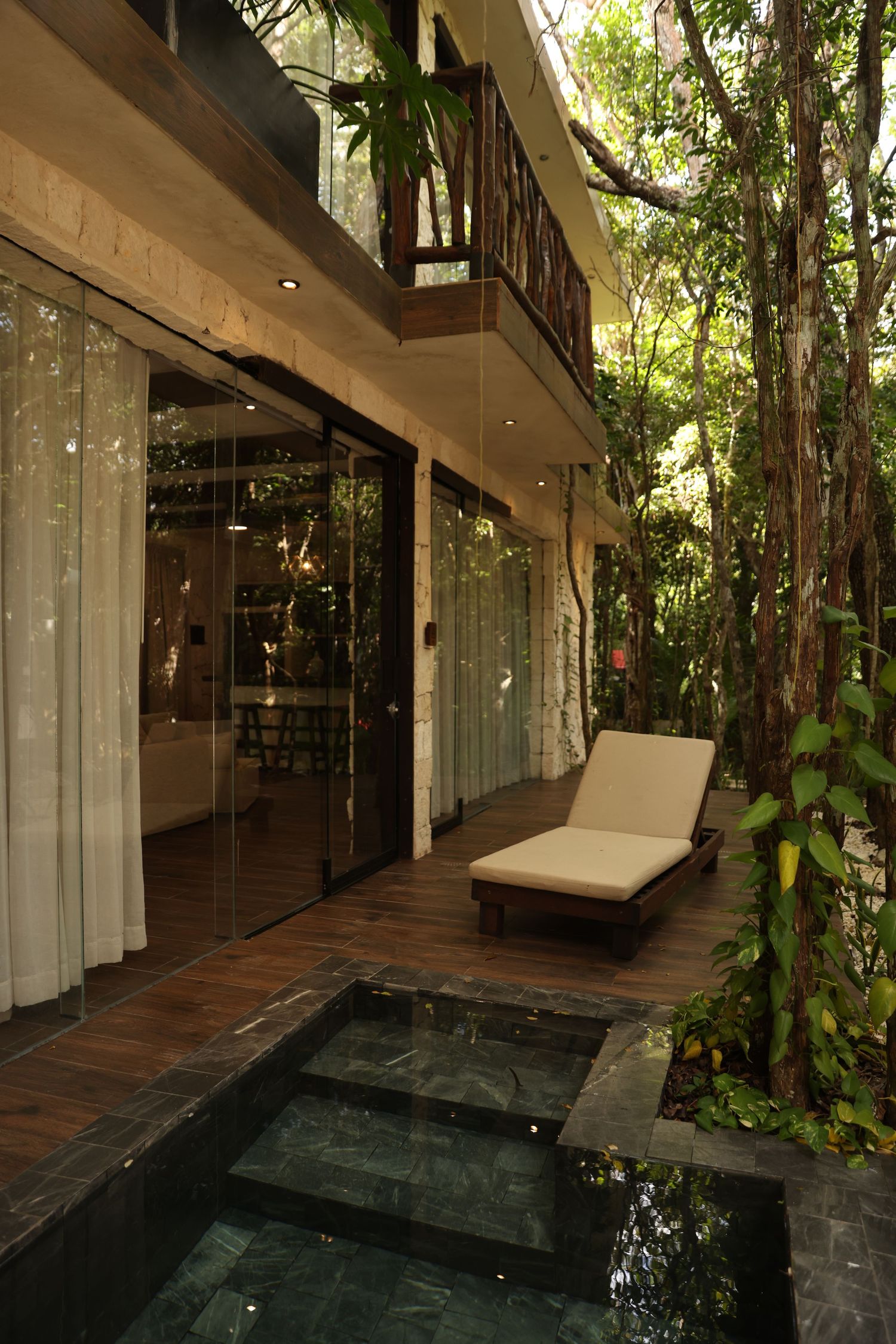 Luxury villa with plunge pool, wooden deck, lounge chair, and lush tropical trees surrounding glass-walled interior.