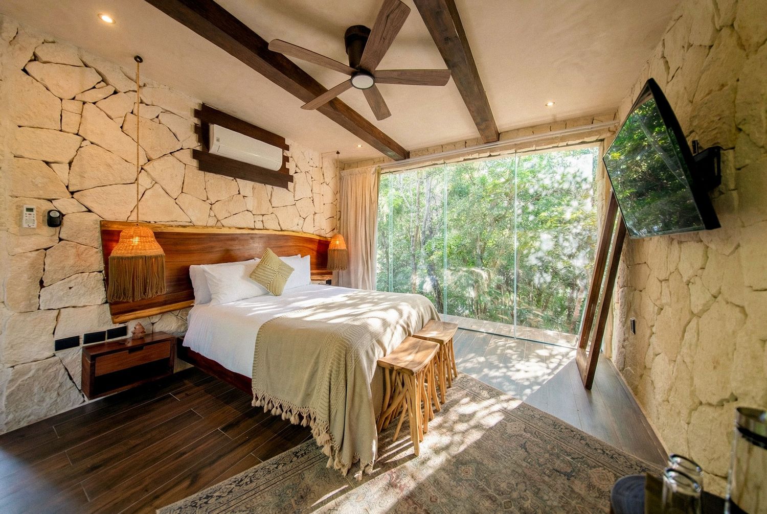 Rustic luxury bedroom with stone walls, wooden beam ceiling, king bed, and large window overlooking lush green forest.