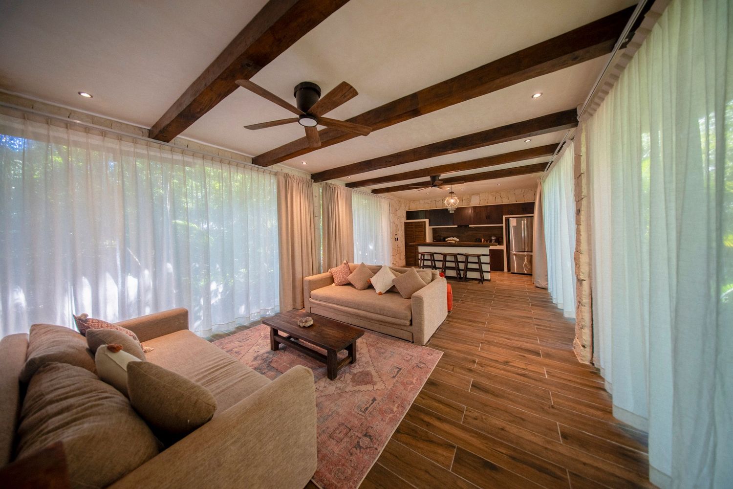 Spacious living room with wood beam ceiling, ceiling fan, sheer curtains, hardwood floors, and open kitchen in background.