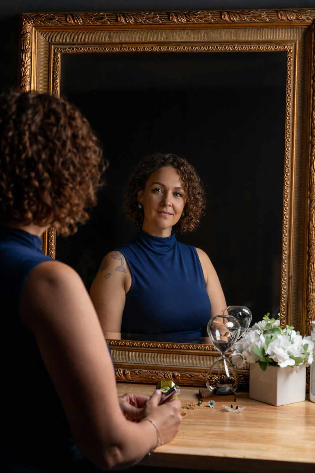 A reflection shows someone sitting at a wooden vanity with an ornate gold mirror and white flowers.