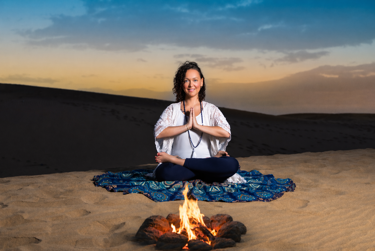 Woman meditating in lotus pose by campfire on desert sand dunes at sunset with hands in prayer position.