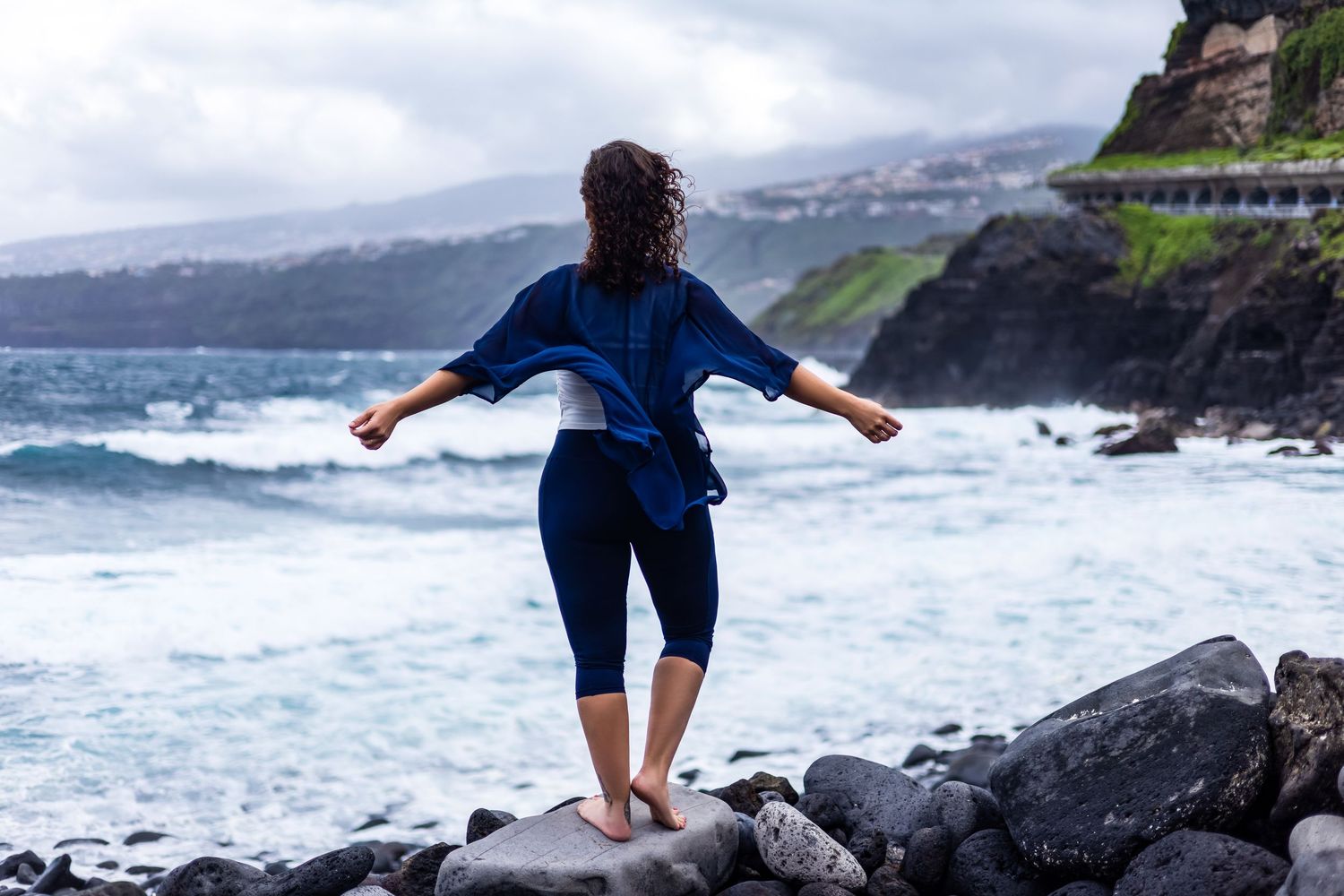 Woman standing barefoot on rocky shore with arms outstretched, facing dramatic ocean waves under overcast skies.
