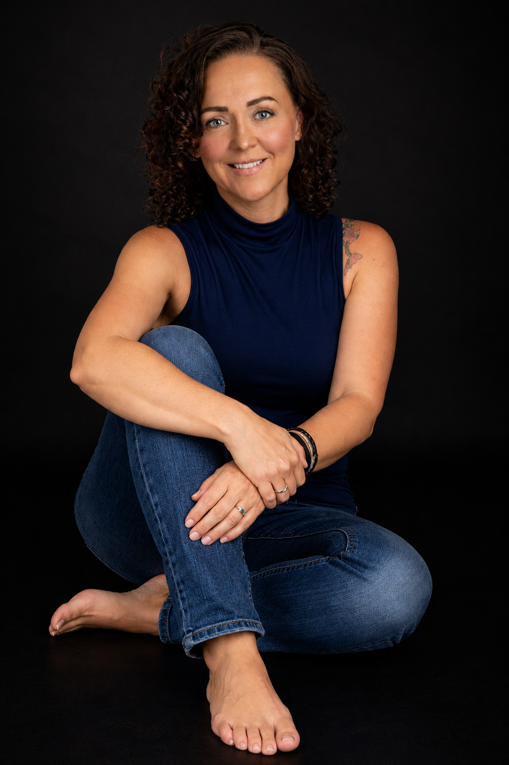 Woman with curly hair sitting cross-legged on floor, wearing blue top and jeans, smiling against dark background.