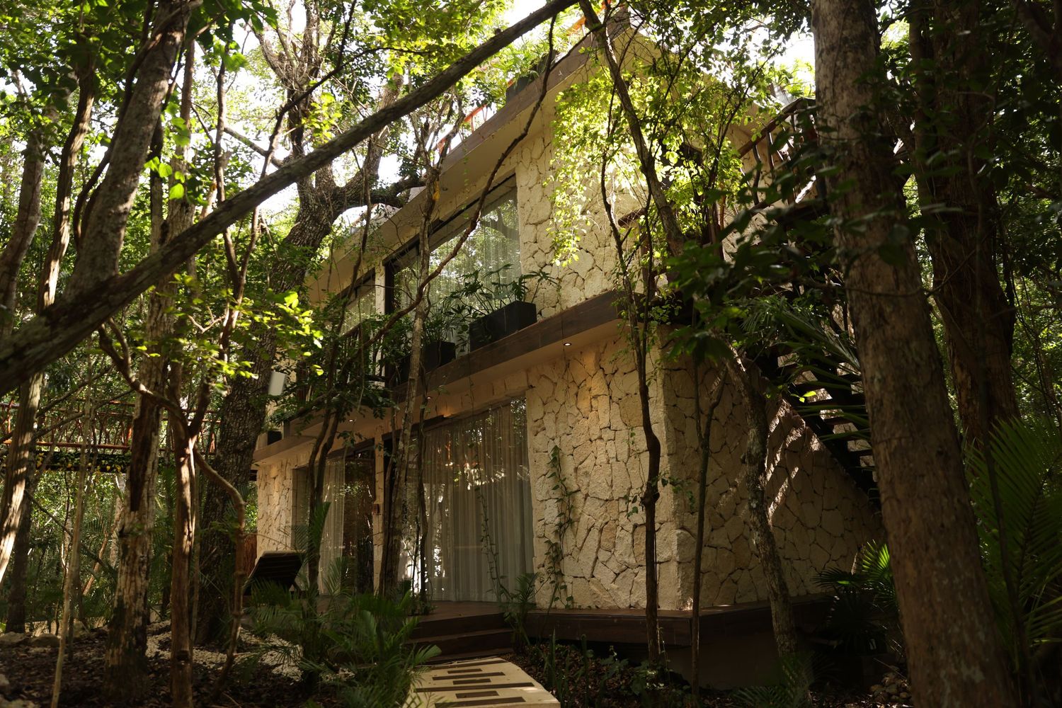 Stone villa nestled among lush tropical trees with wooden walkway and climbing vines in jungle setting.