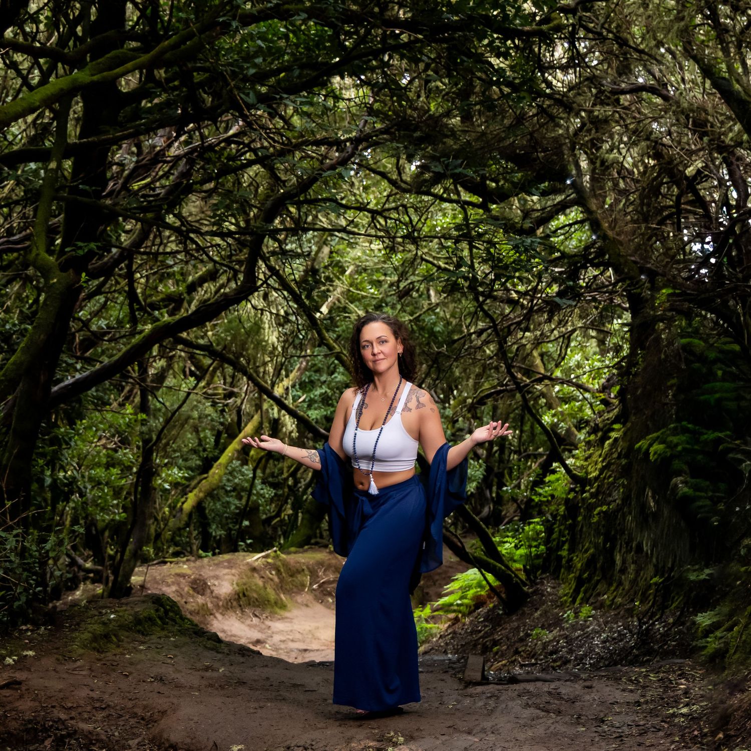 Woman in blue skirt and white top posing gracefully on a forest path surrounded by lush green trees.