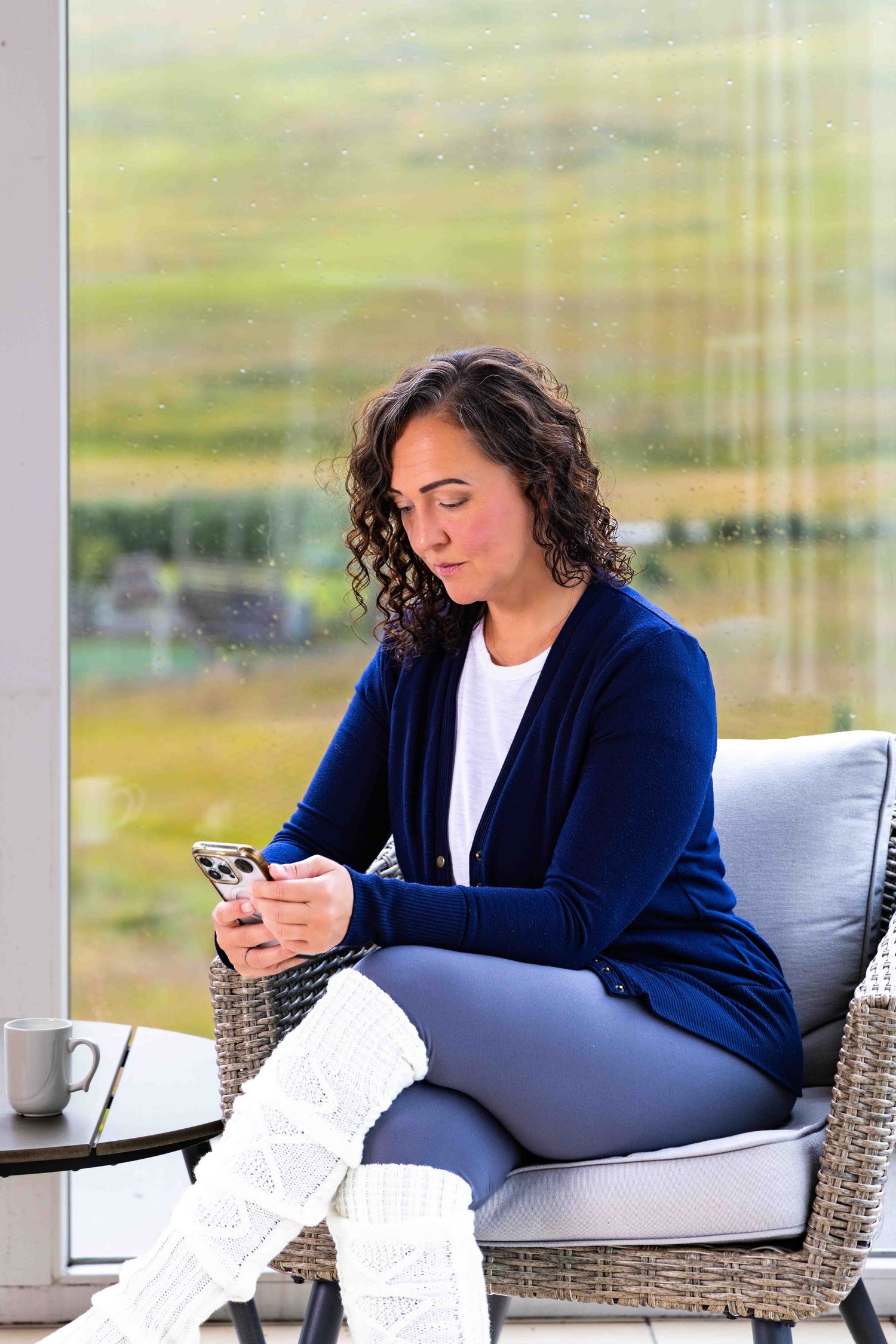 Woman in blue cardigan sitting in wicker chair using smartphone near large window with green landscape.