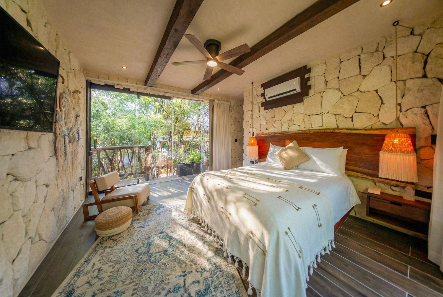 Rustic bedroom with stone walls, wooden beams, ceiling fan, lush garden view, and a white-draped bed with warm lighting.