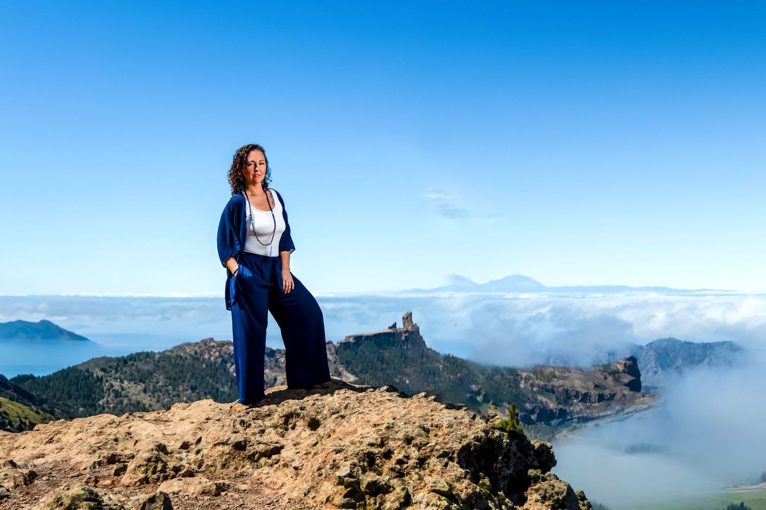 Woman in blue outfit standing on rocky mountain peak with clouds and volcanic landscape below under clear blue sky.