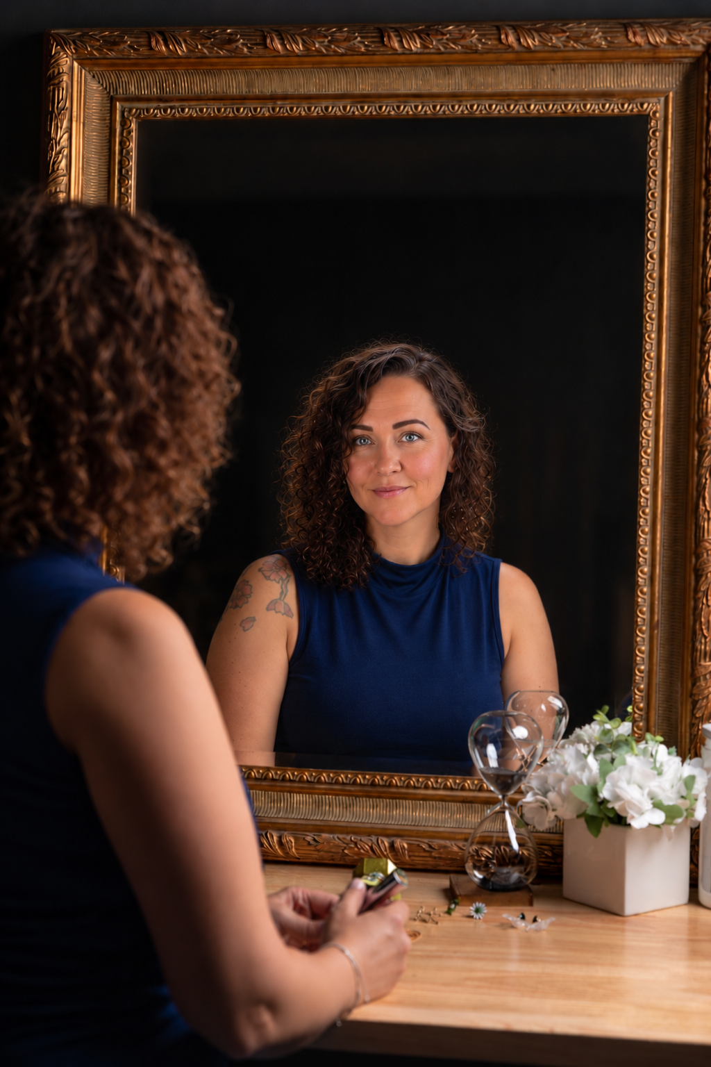Woman with curly hair in blue top gazes at her reflection in an ornate gold-framed mirror on a wooden table.