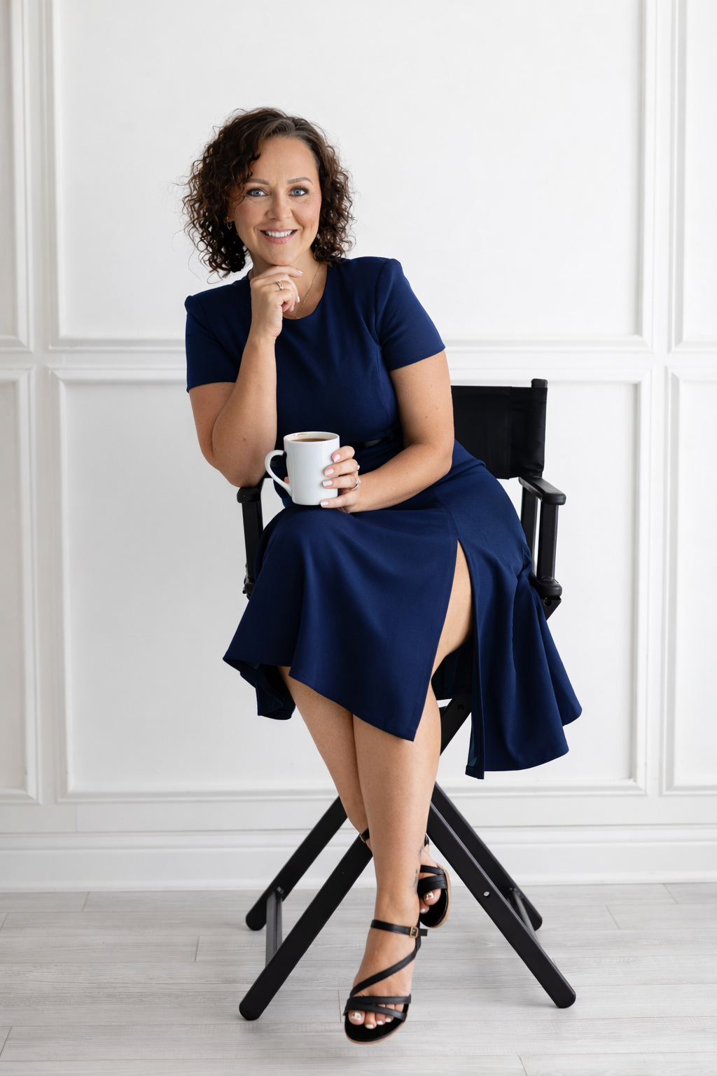 A person in a navy dress sits casually in a white director's chair against a bright studio backdrop.