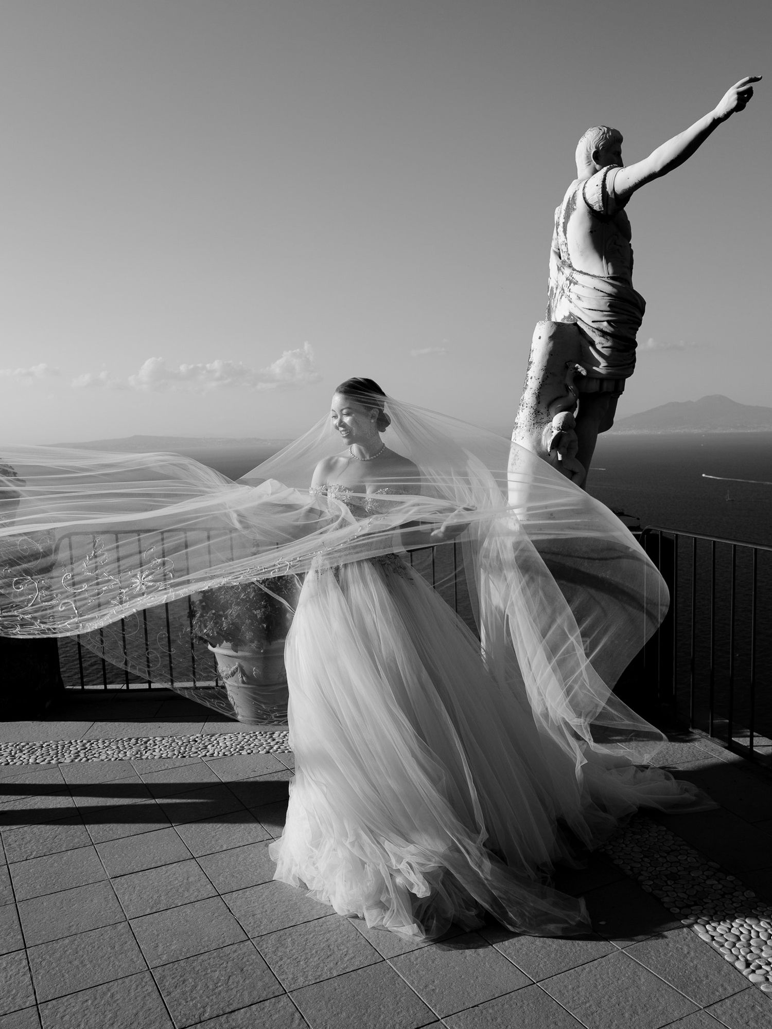Flowing white wedding dress veil dances in the wind against a dramatic coastal vista in black and white photography.