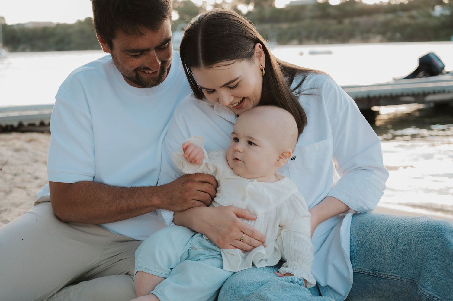 Happy young family with baby sitting on beach, parents smiling at infant in white outfit near waterfront.