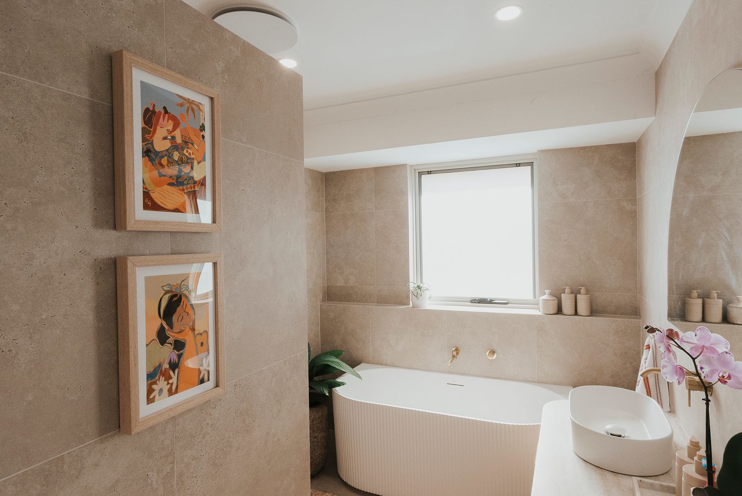 Modern luxury bathroom with freestanding white soaking tub, beige tile walls, framed artwork, and fresh orchid flowers.