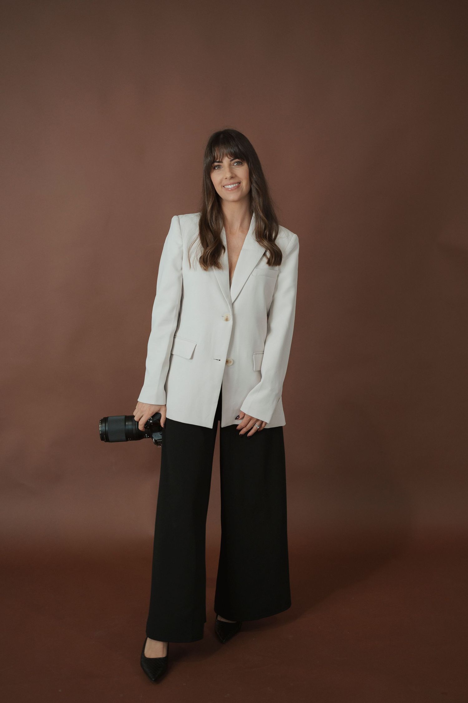 A woman in a white blazer and black wide-leg pants holds a camera, posing confidently against a brown background.