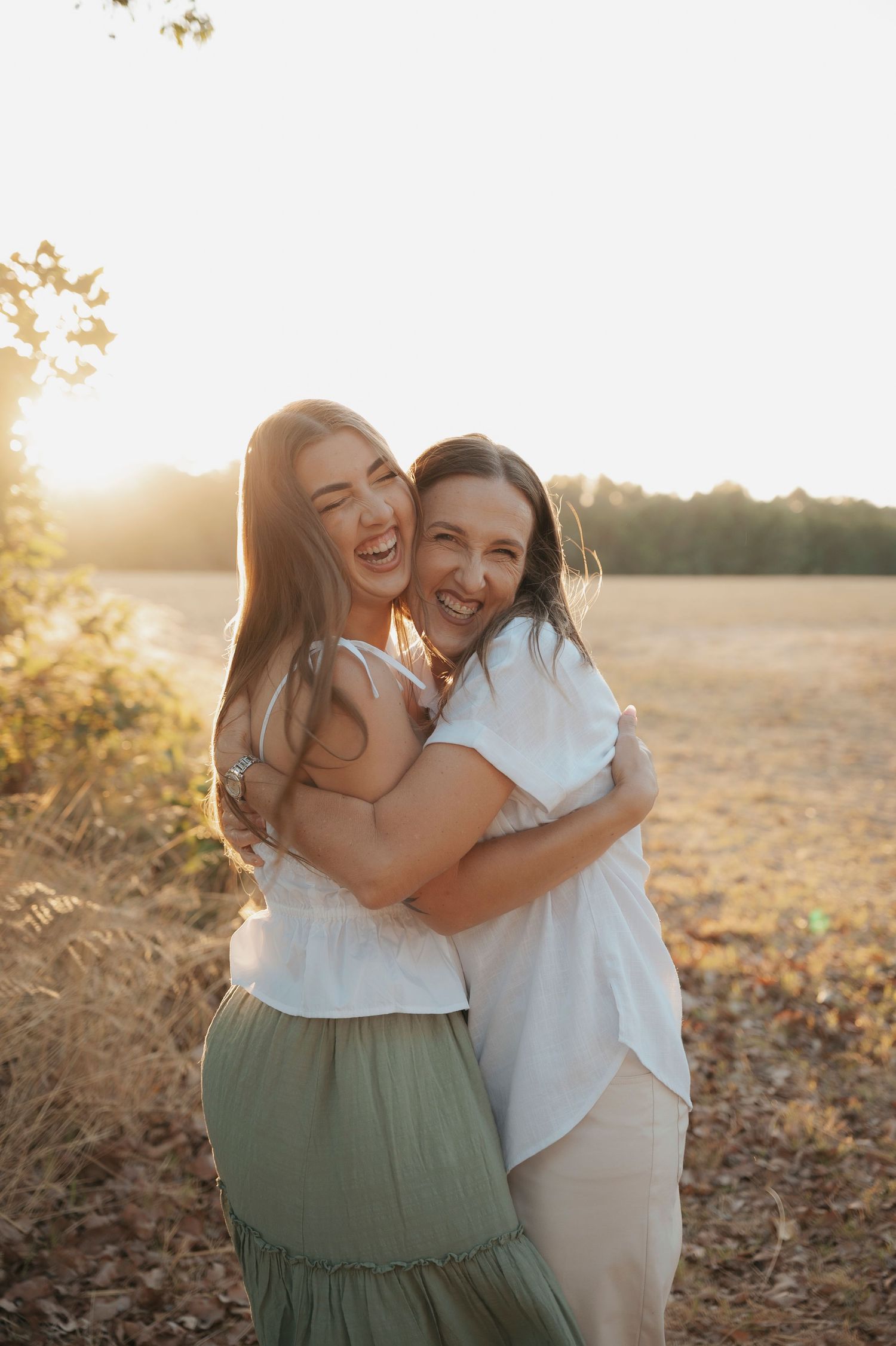 Two women hugging and laughing in a golden sunlit field at sunset, wearing white and green outfits.