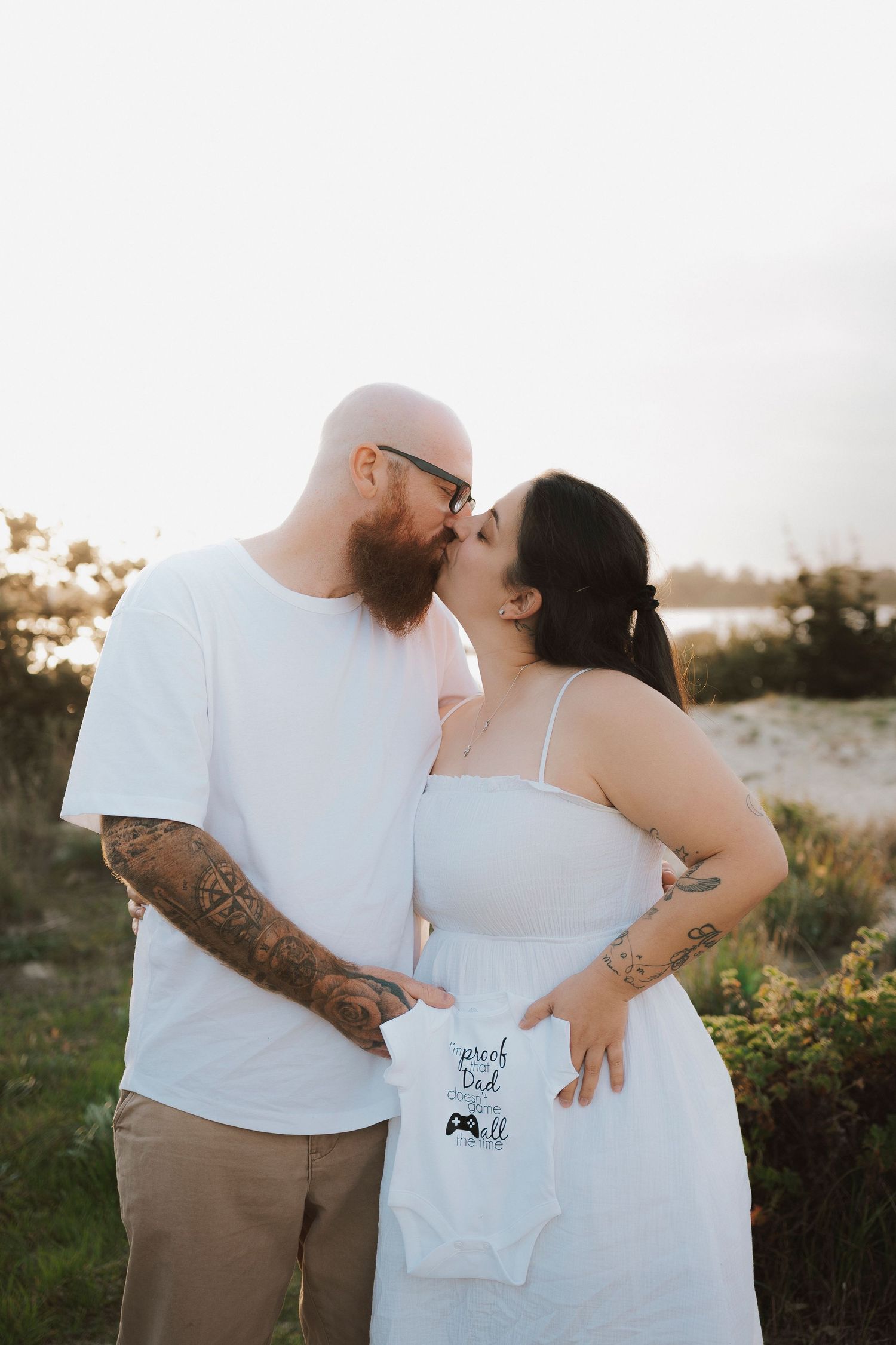 Couple kissing outdoors at sunset, holding a white baby onesie, announcing pregnancy, with tattoos and white outfits.