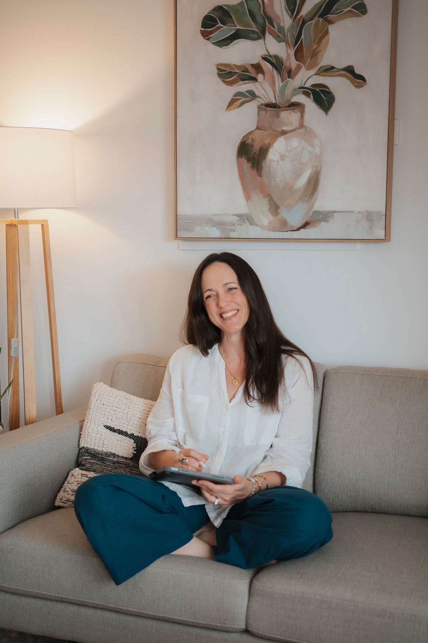 Woman smiling on gray sofa holding tablet, wearing white blouse and teal pants, with plant painting on wall.
