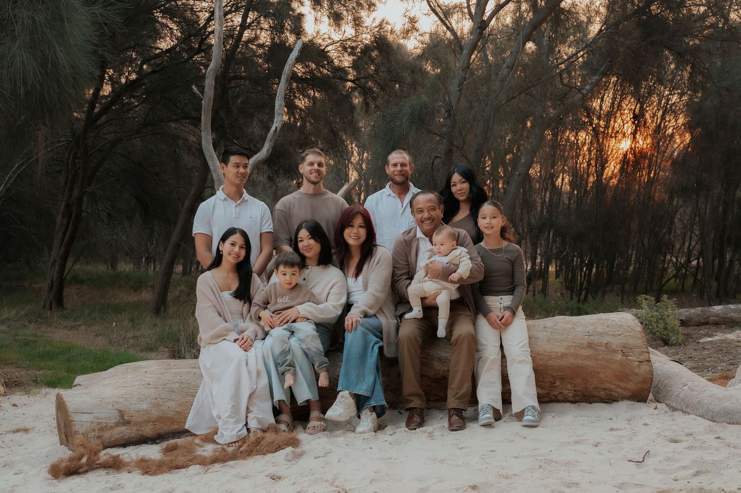 Large family portrait at sunset in a forest clearing, seated on a log in neutral tones.
