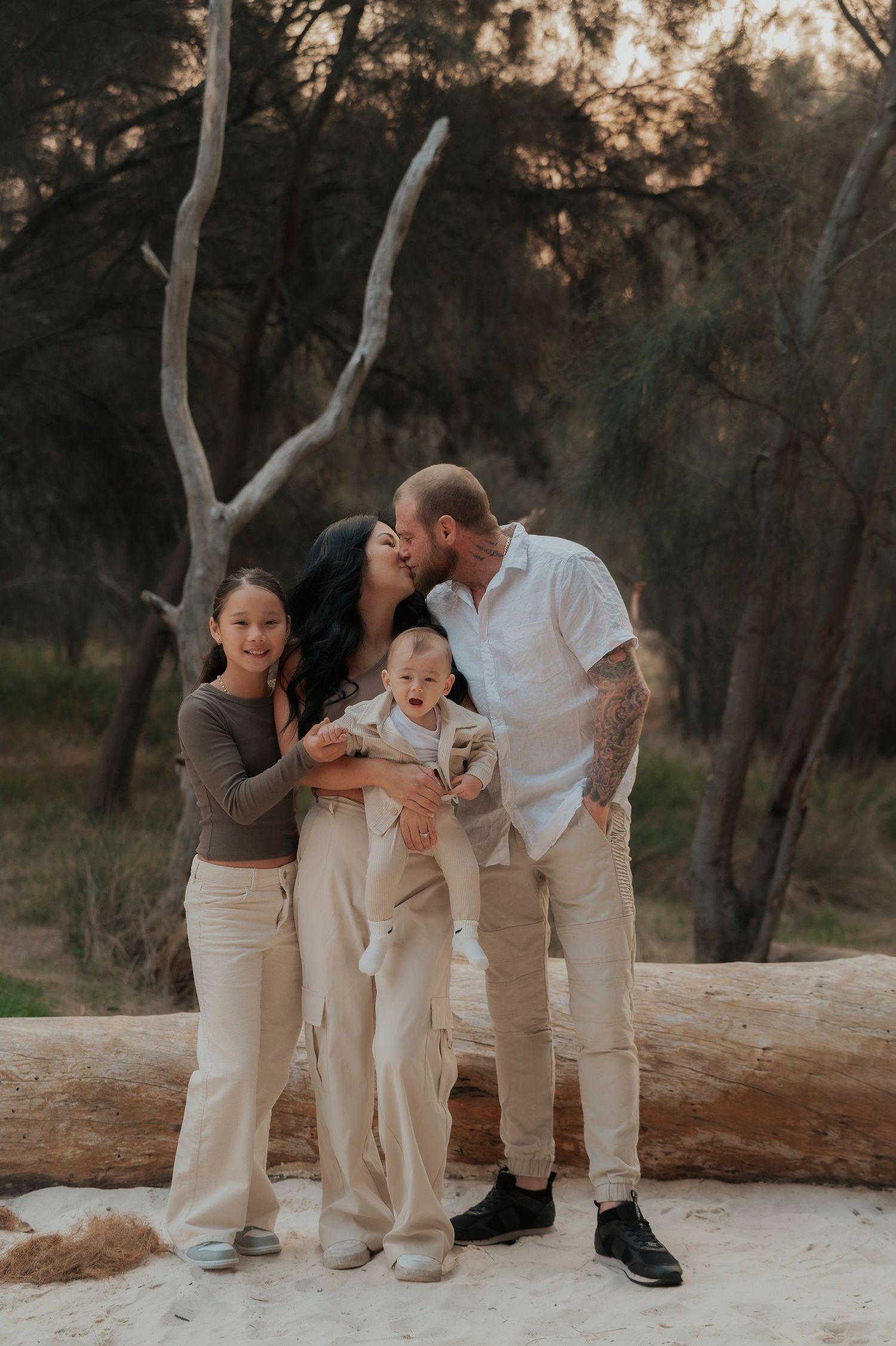 Family of four posing outdoors among trees, parents kissing while holding a laughing baby, with a young girl beside them.
