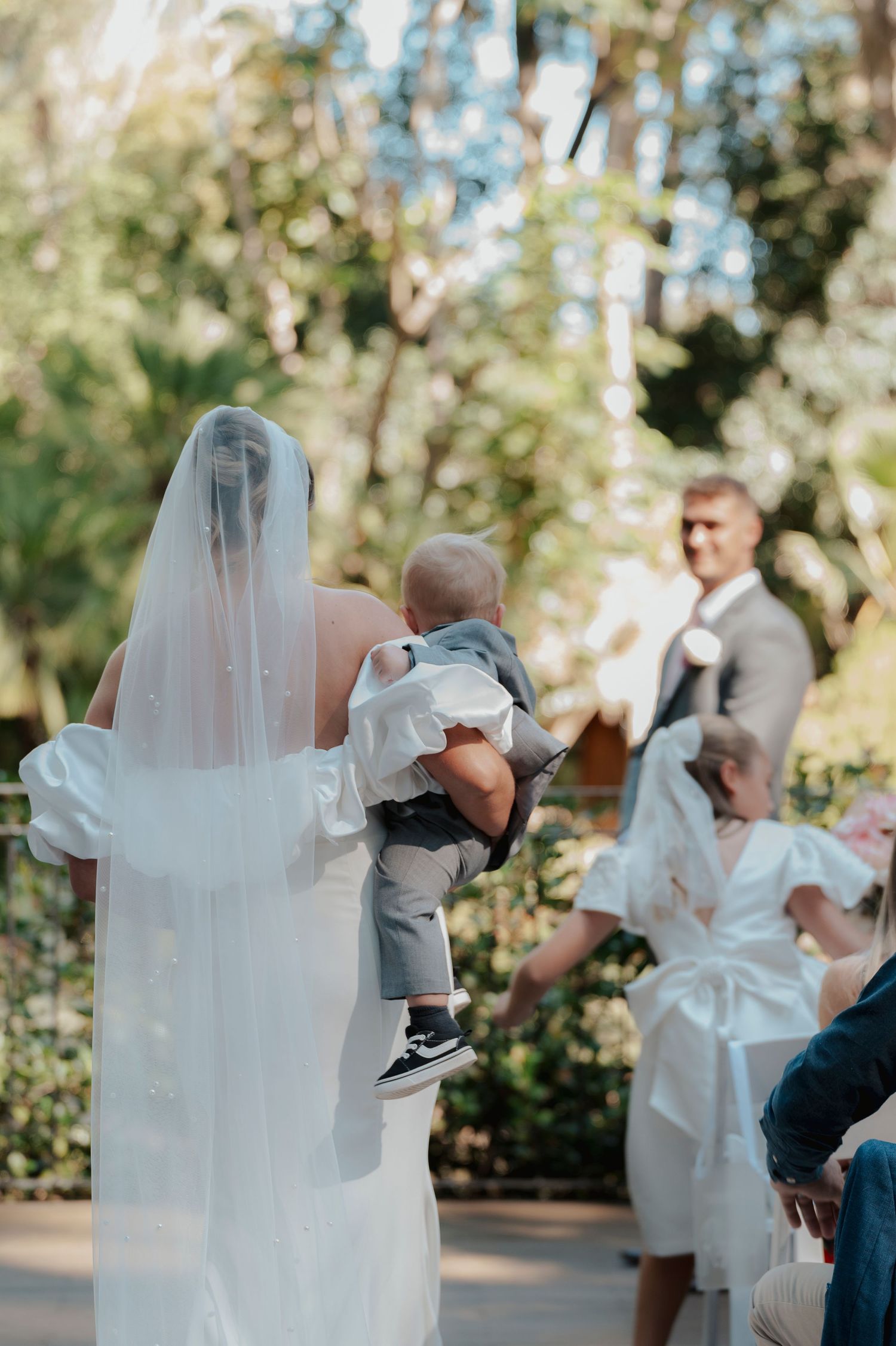 Bride in white gown and veil walks down outdoor aisle carrying baby toward groom during wedding ceremony.