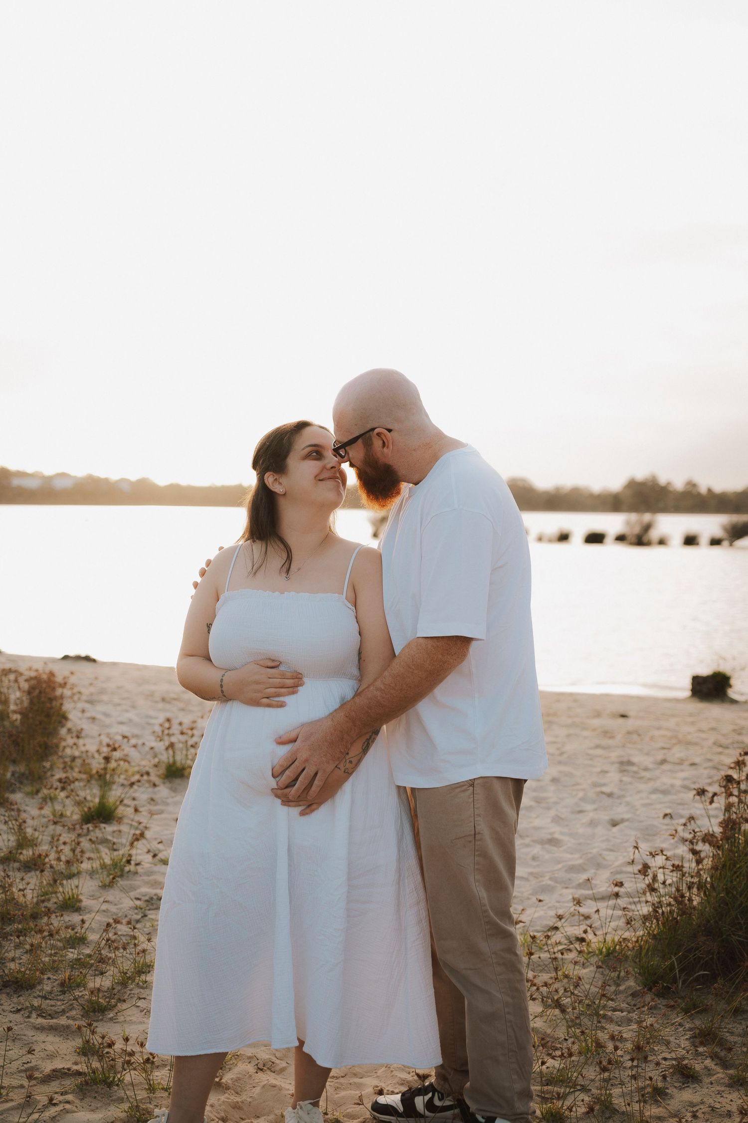 Couple sharing a tender moment on a sandy beach at sunset, woman in white dress cradling baby bump.