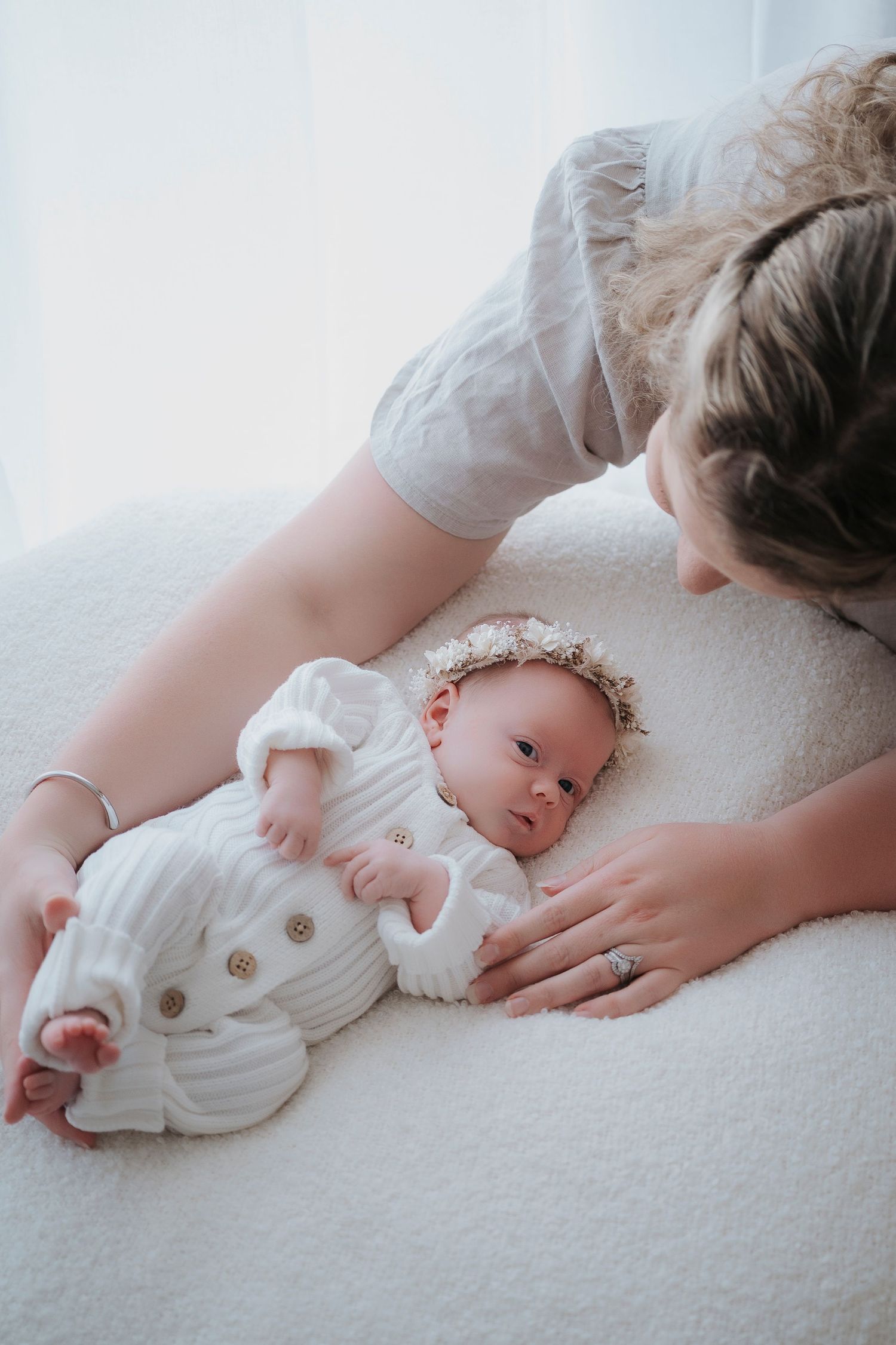 Mother leaning over newborn baby in white outfit and floral headband lying on soft white surface.