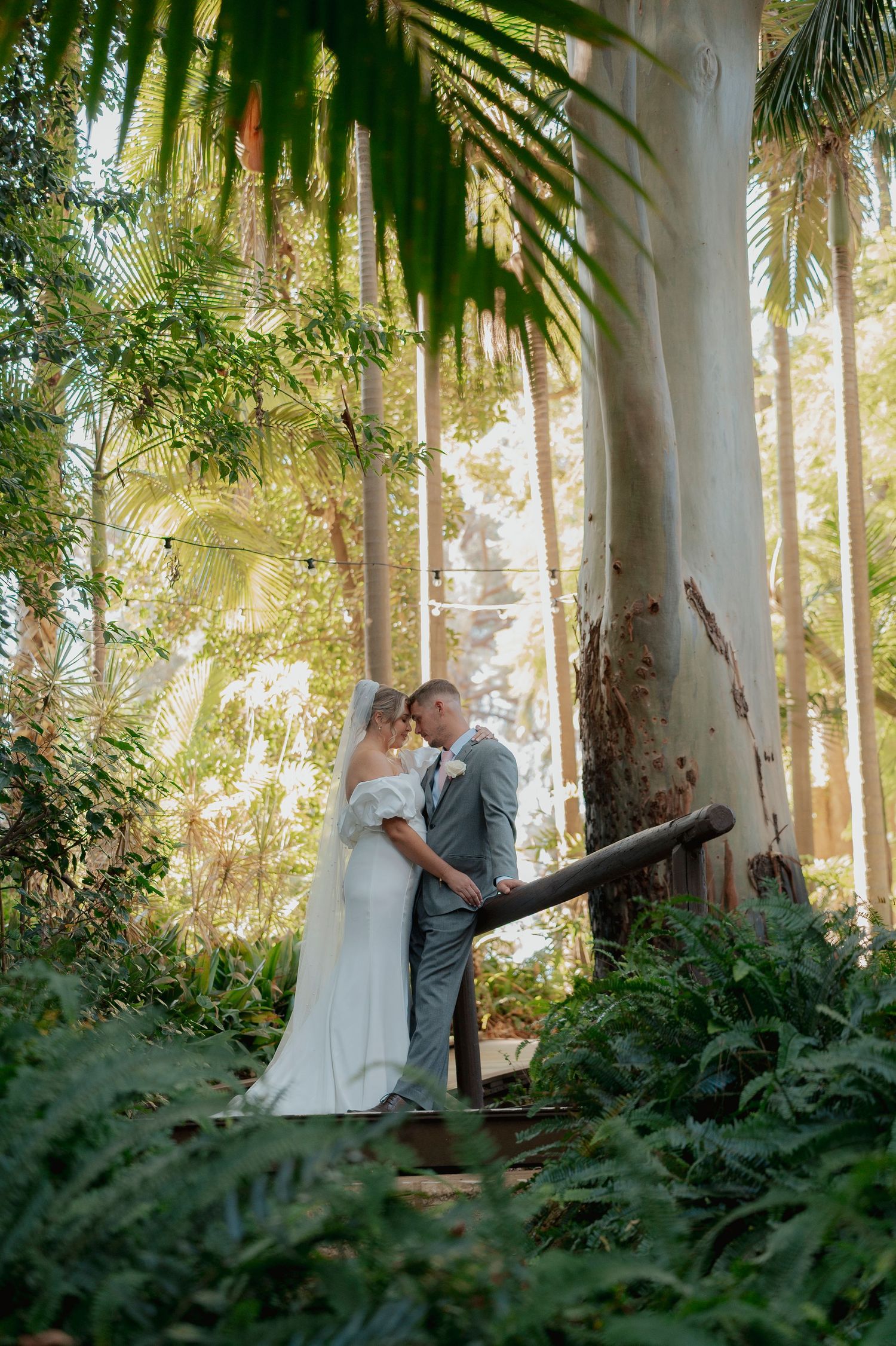 Bride and groom share a romantic moment surrounded by lush tropical greenery and tall trees in a garden setting.