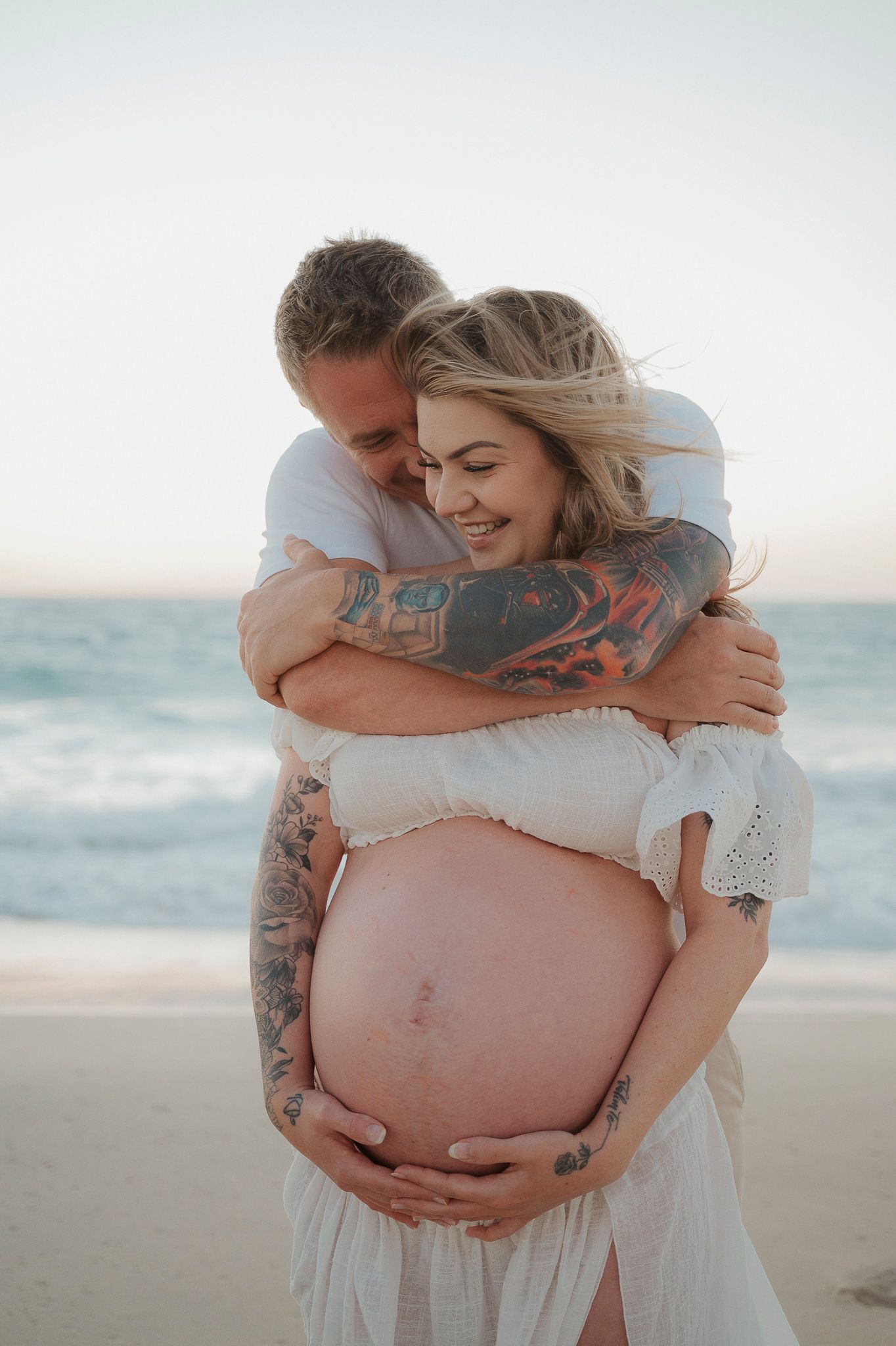 Couple embracing on beach at sunset, pregnant woman in white dress holding baby bump, man with tattoos hugging her from behind.