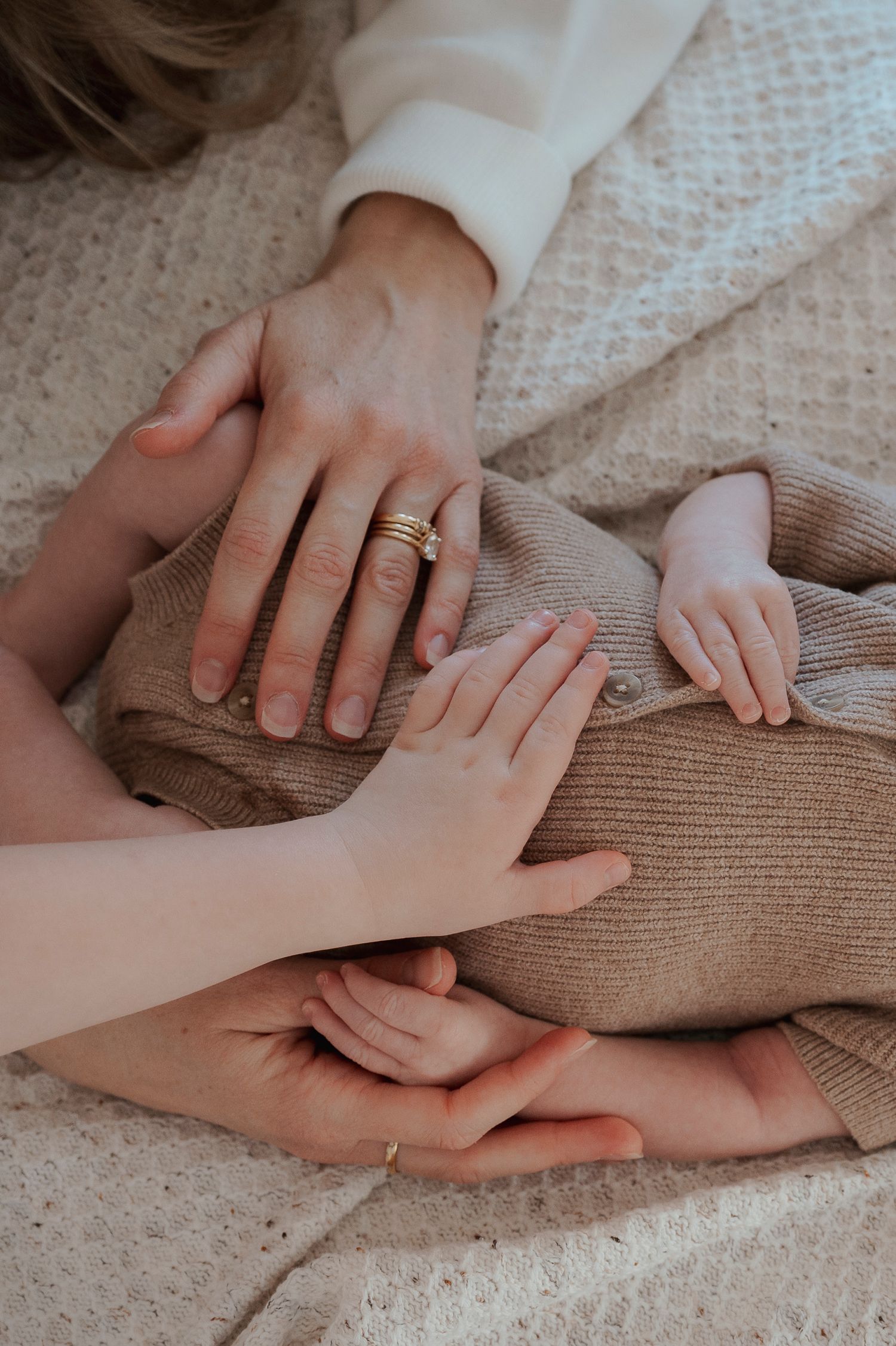 Family hands cradling a newborn baby in a beige knit outfit on a soft blanket, mother wearing gold rings.