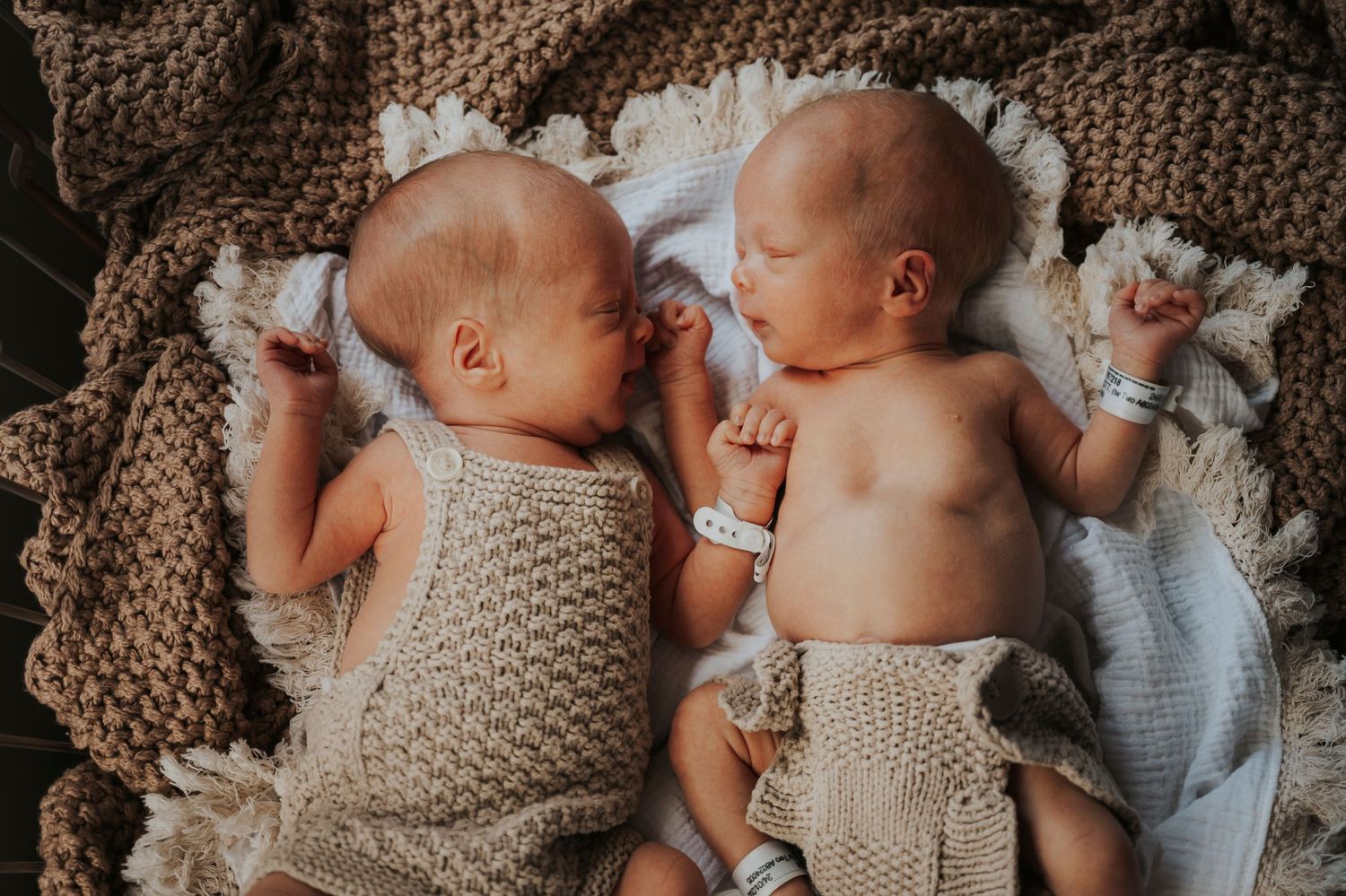 Two newborn twins lying side by side on a knitted blanket, wearing crochet outfits with hospital wristbands.