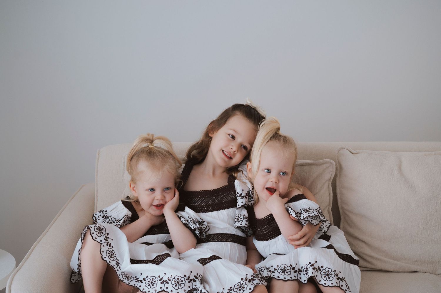 Three young girls in matching floral dresses sitting together on a cream sofa, smiling for a family portrait photo.