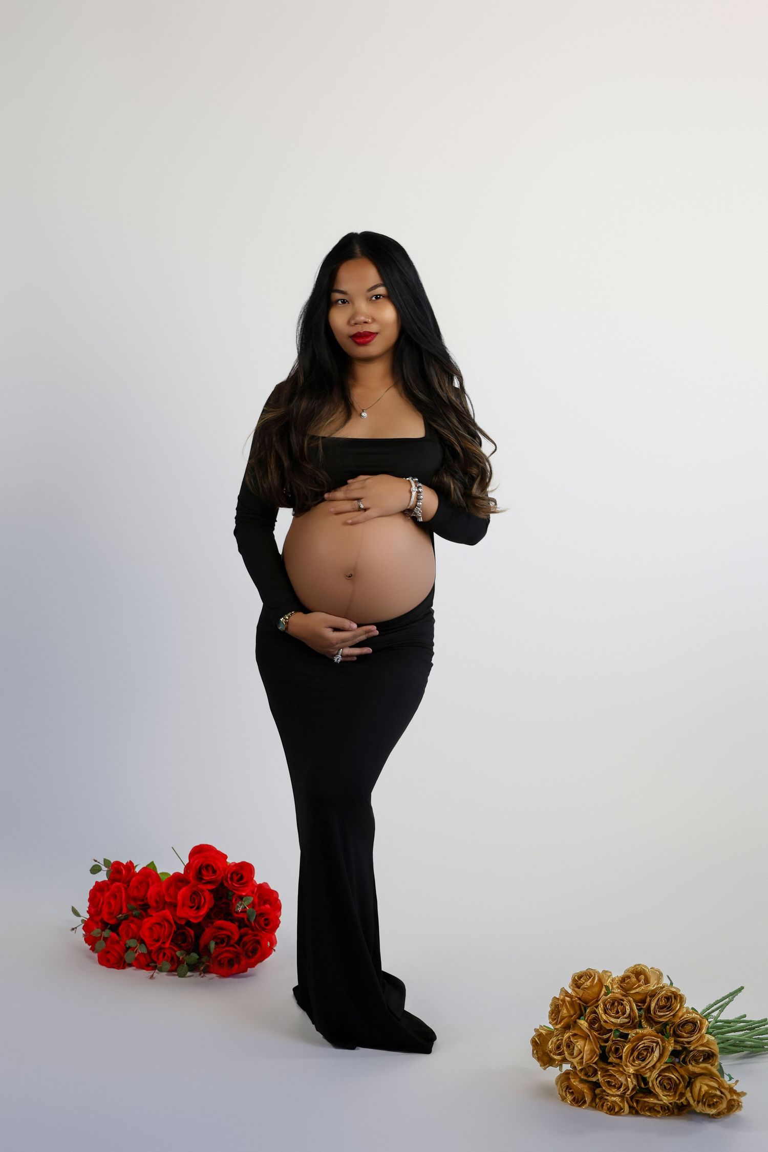 Maternity photo featuring a woman in black dress posing with red and gold rose bouquets against white backdrop.