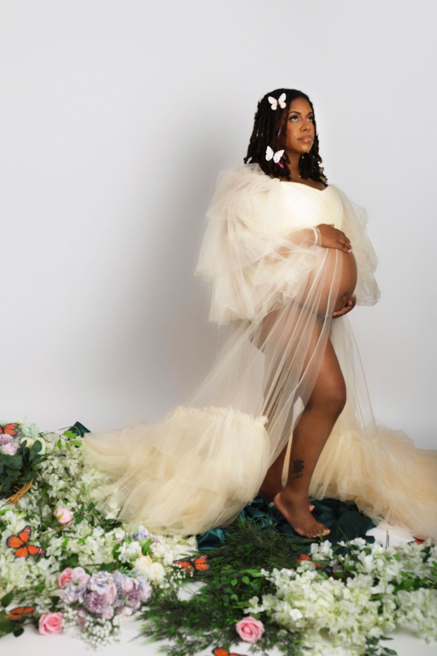 Ethereal maternity photoshoot featuring flowy tulle gown amidst romantic floral arrangements on white backdrop.