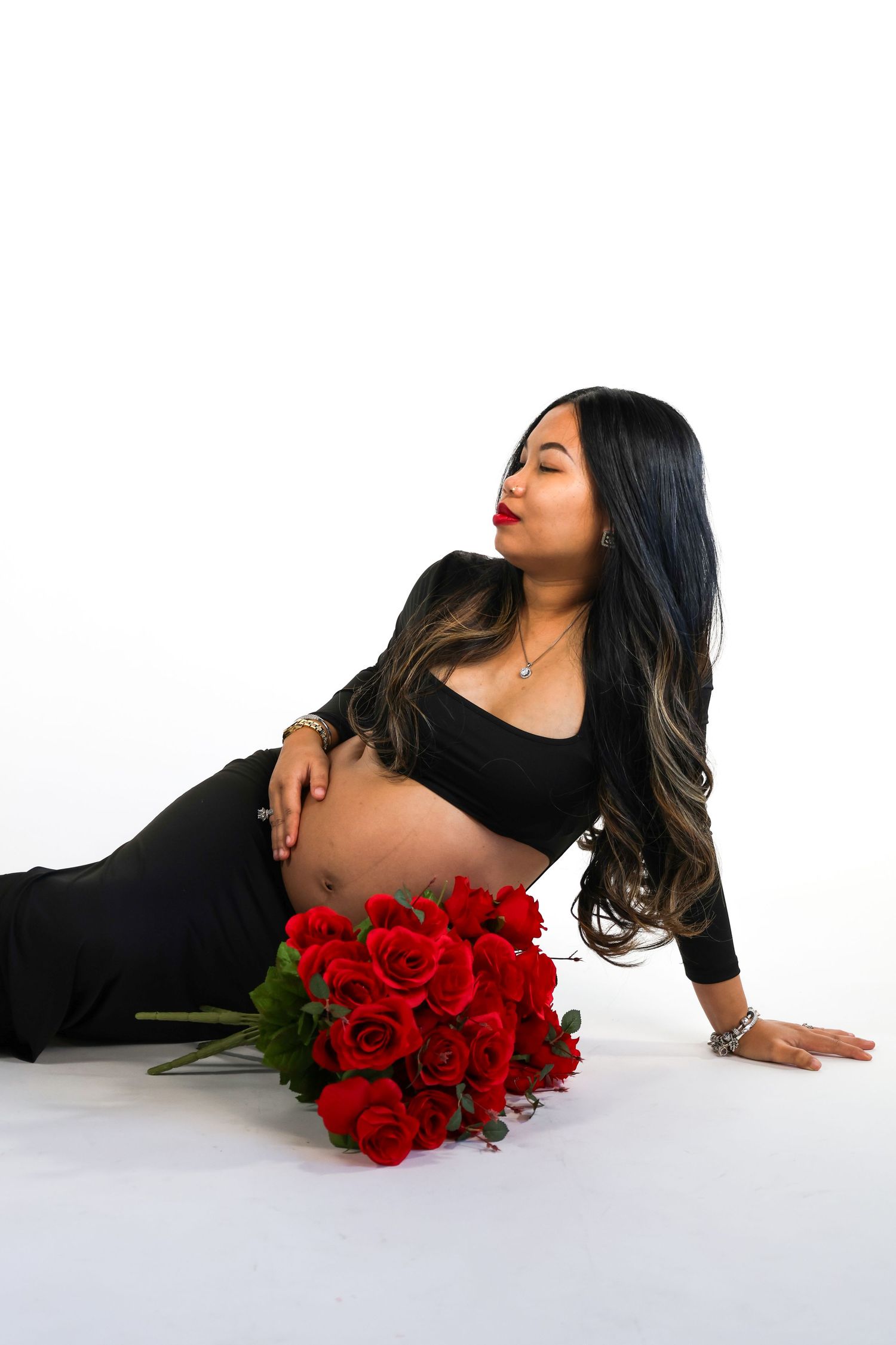 A maternity photo featuring an elegant black dress, long flowing hair, and a vibrant bouquet of red roses on white background.